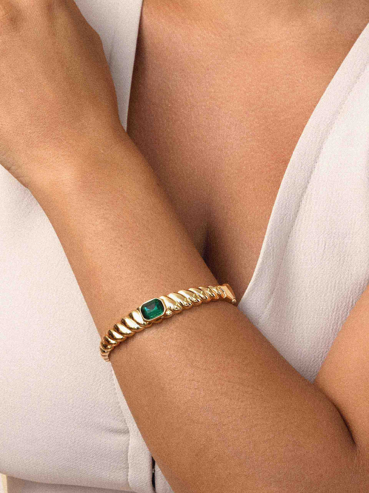 Close-up of a person wearing a gold chain bracelet with a green gemstone, crossed over white clothing; warm, elegant, understated luxury.