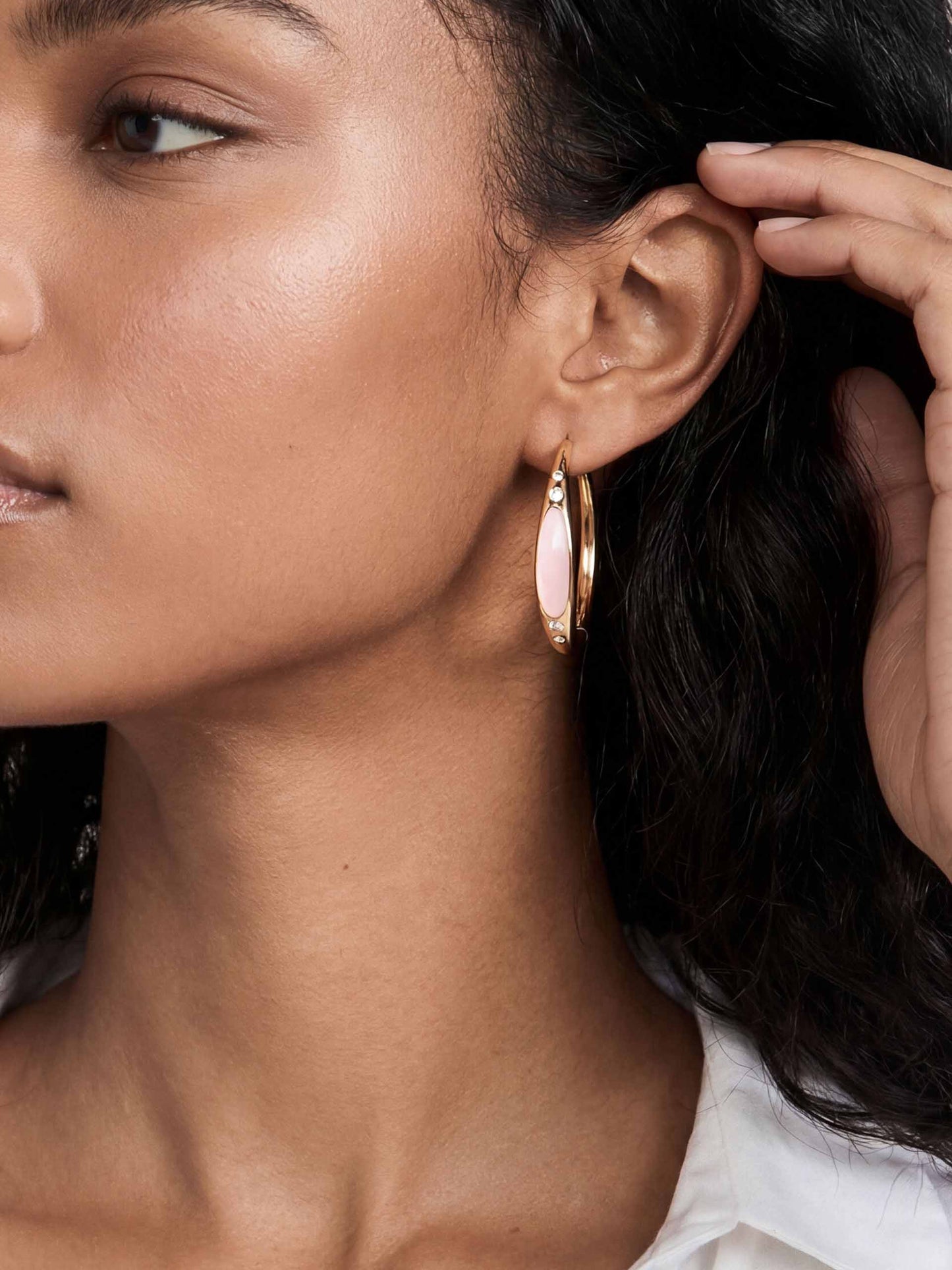 Close-up of an ear wearing a gold hoop earring with a smooth pink gemstone and three small diamonds. The style is elegant and modern.
