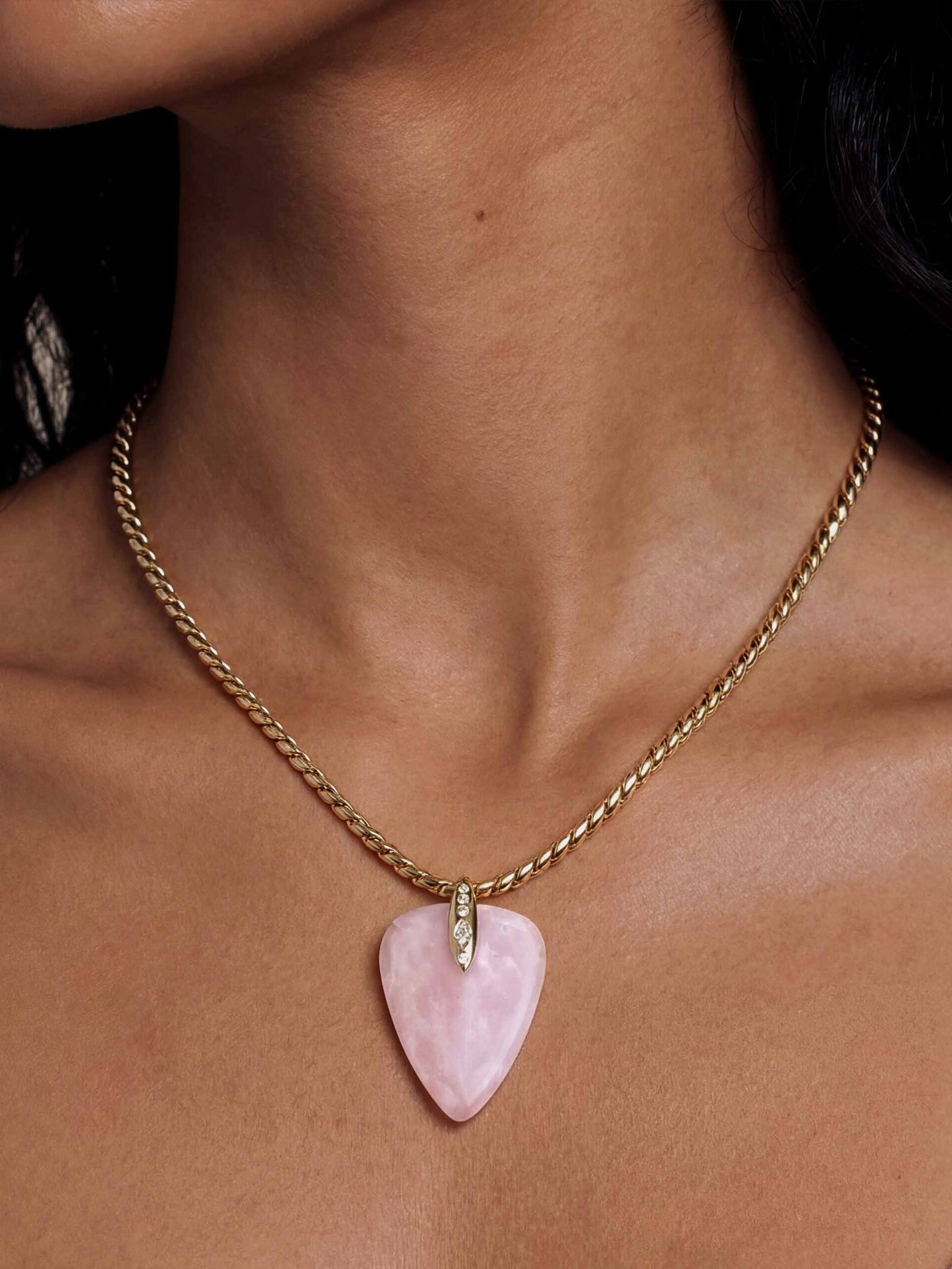 A gold chain necklace with a pink heart-shaped pendant is worn around a woman's neck. The pendant has a small diamond accent on top, set against a bare skin backdrop, exuding elegance.