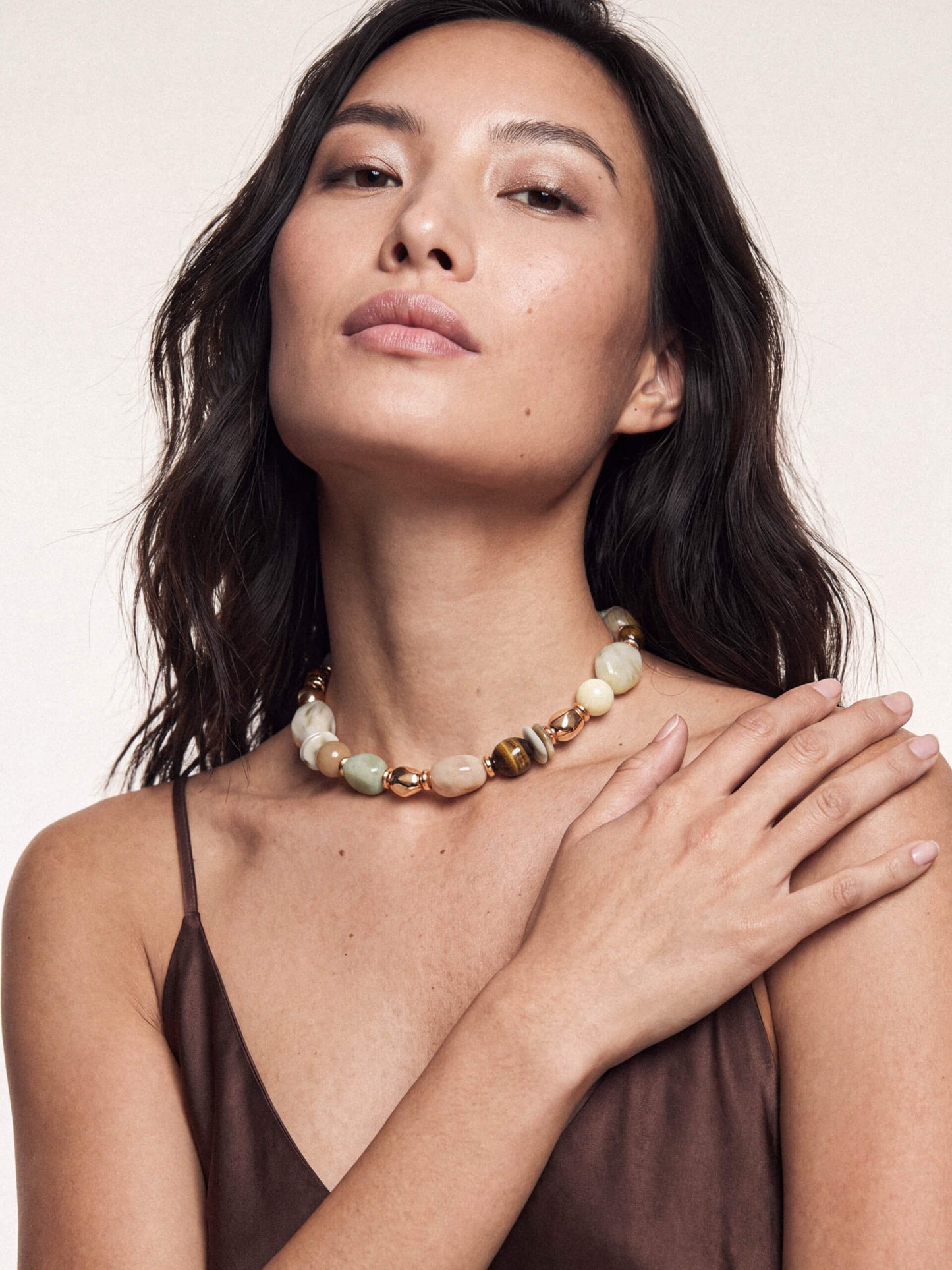 A woman wearing a chunky beaded necklace with natural stone beads in earthy tones of beige, green, and brown, styled with gold spacer beads. She is wearing a brown camisole against a plain, light beige background.
