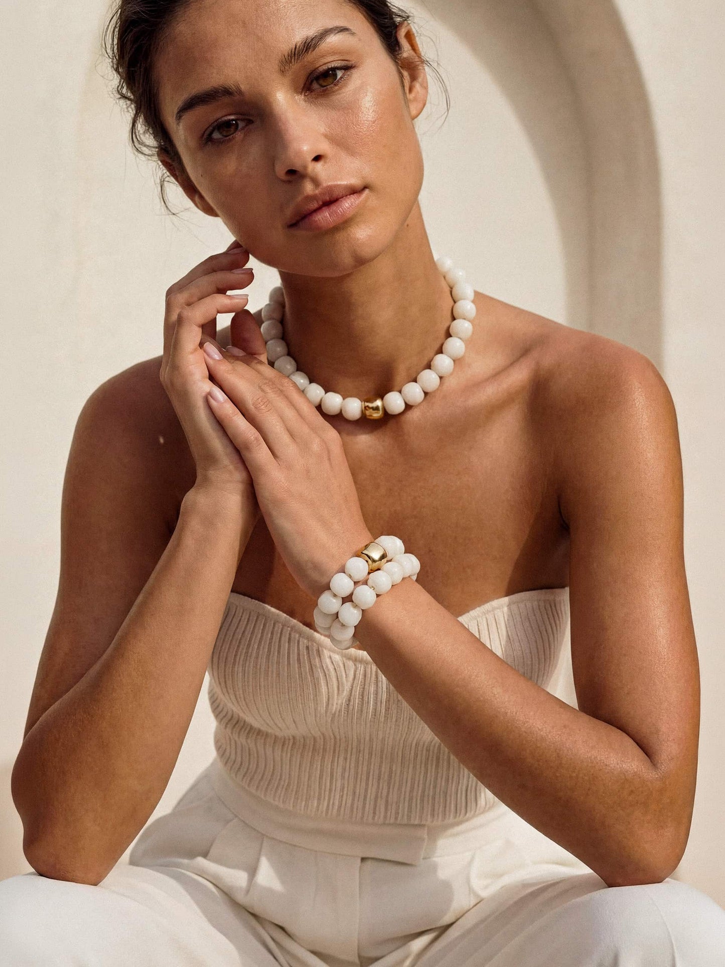 Model in neutral outfit wearing pearl necklace and bracelets, posing with hands near face.