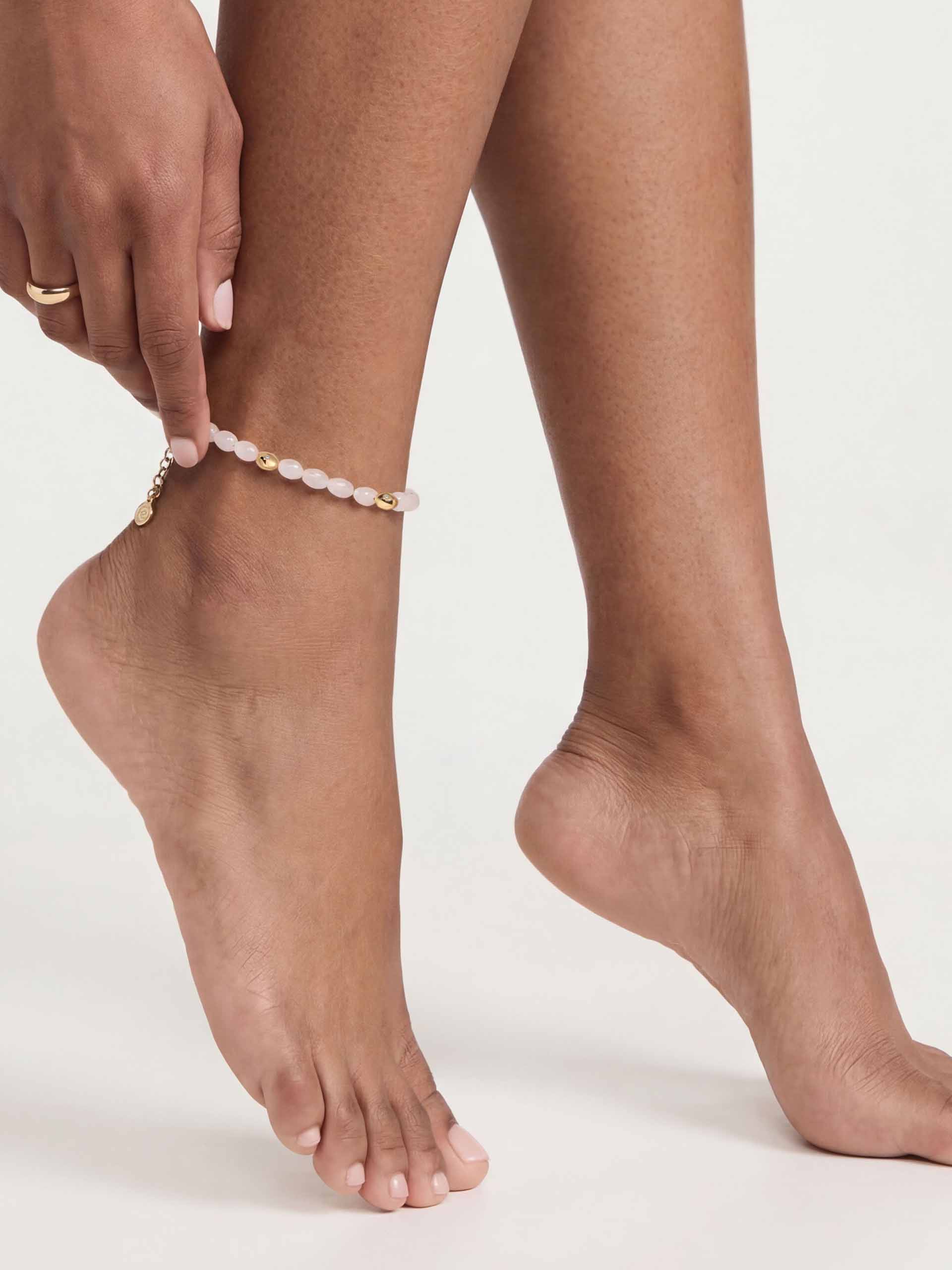 A close-up of an ankle adorned with a delicate gold anklet, featuring soft pink beads and a small gold charm. The skin tone is warm against a white background.