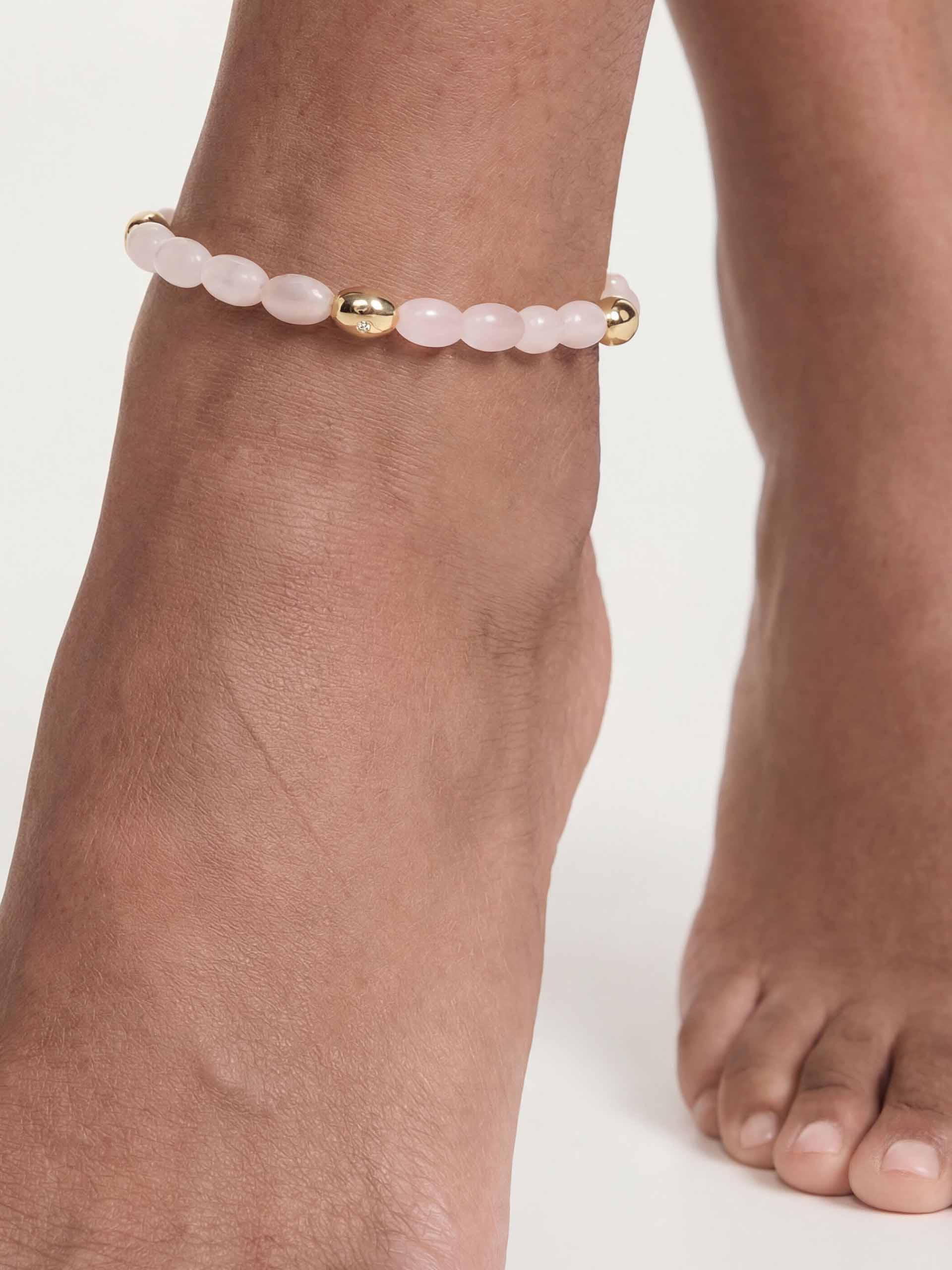 A close-up of an ankle adorned with a delicate gold anklet, featuring soft pink beads and a small gold charm. The skin tone is warm against a white background.