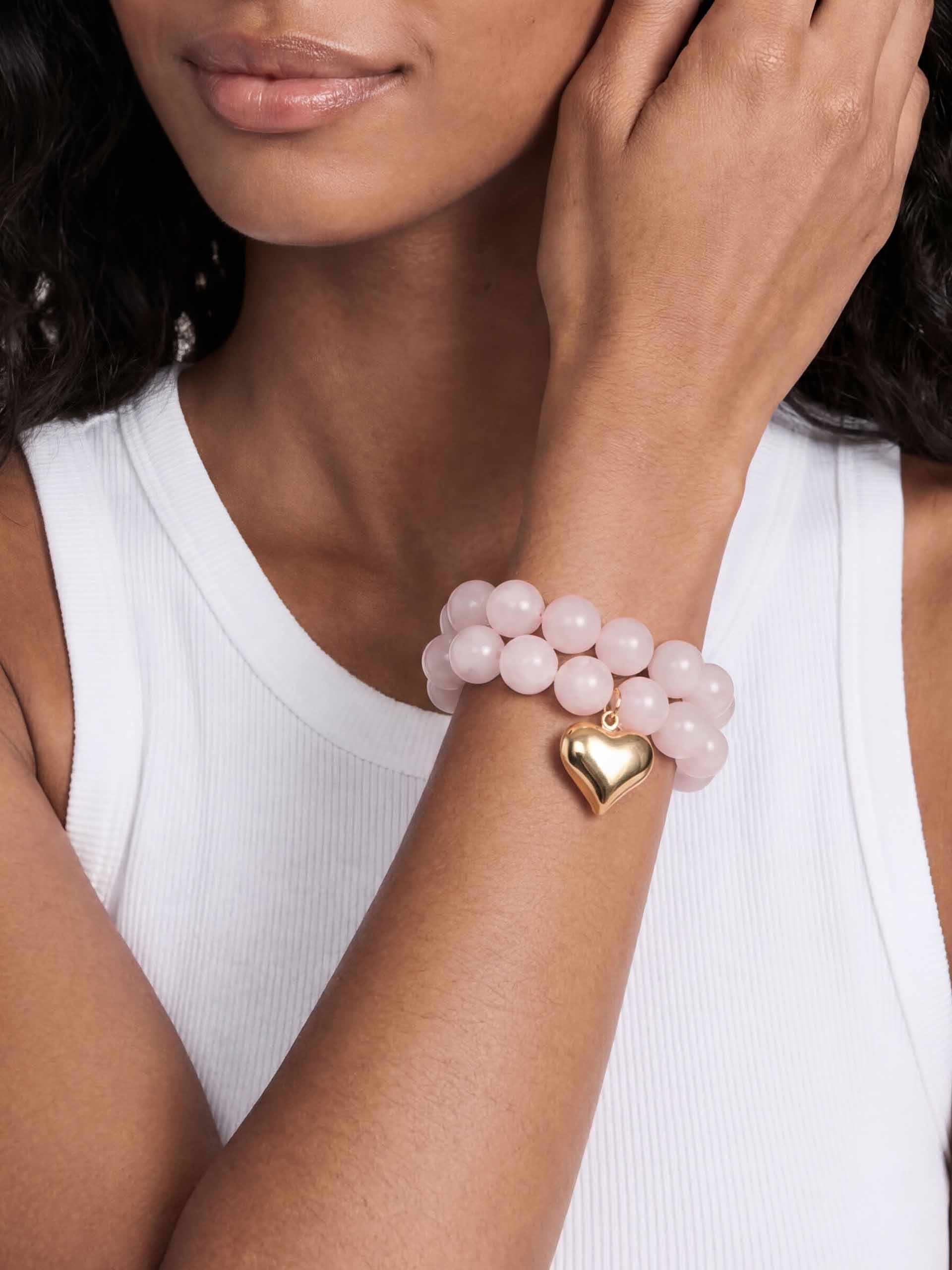 A person wearing a white tank top displays a bracelet of pink beads with a gold heart charm, evoking elegance and simplicity.