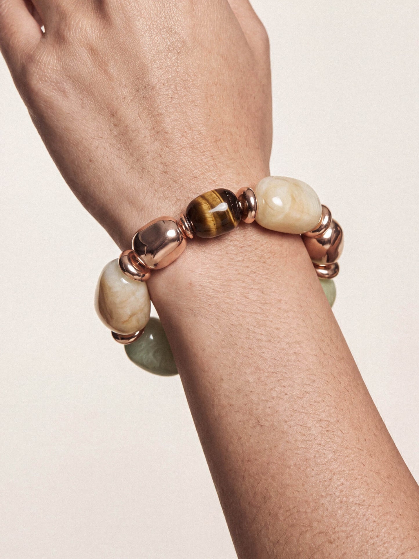 A close-up of a person's wrist wearing a beaded stretch bracelet with polished rose gold metal beads, cream-colored stone beads, and a central brown tiger's eye bead.