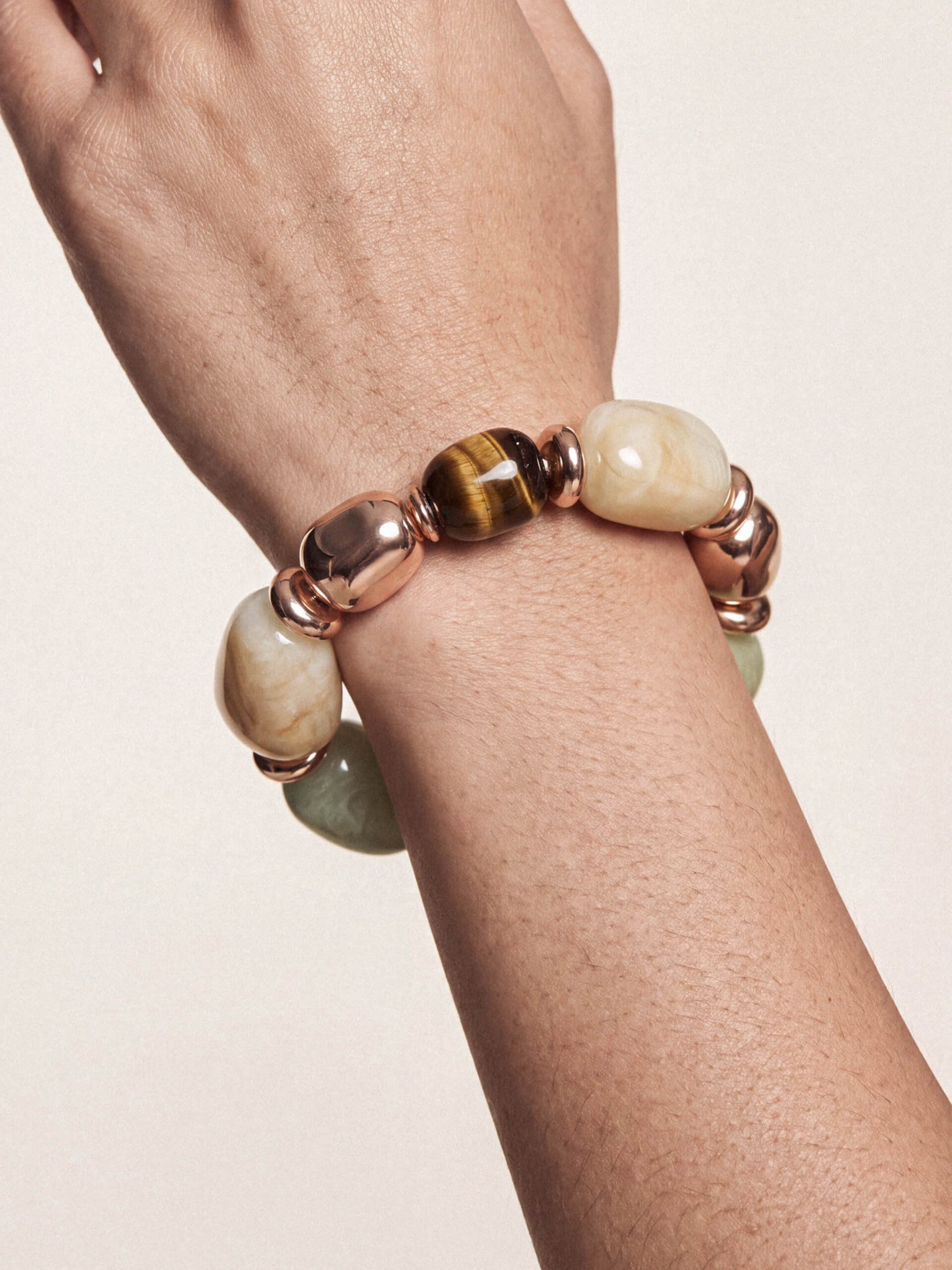 A close-up of a person's wrist wearing a beaded stretch bracelet with polished rose gold metal beads, cream-colored stone beads, and a central brown tiger's eye bead.