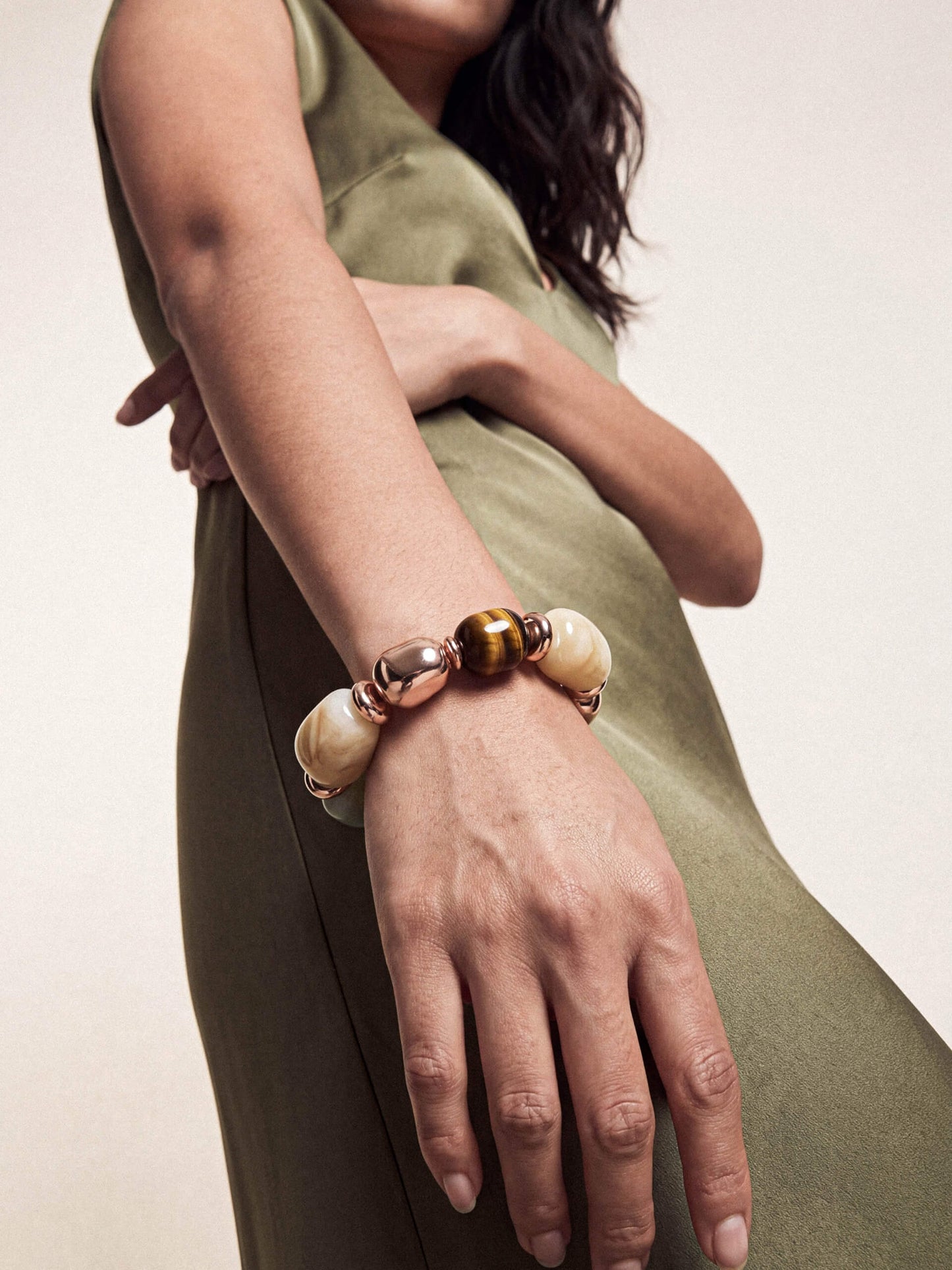 A close-up of a woman's wrist wearing a beaded bracelet with cream-colored round beads, a metallic rose gold bead, and a tiger's eye accent bead, set against an olive green dress.