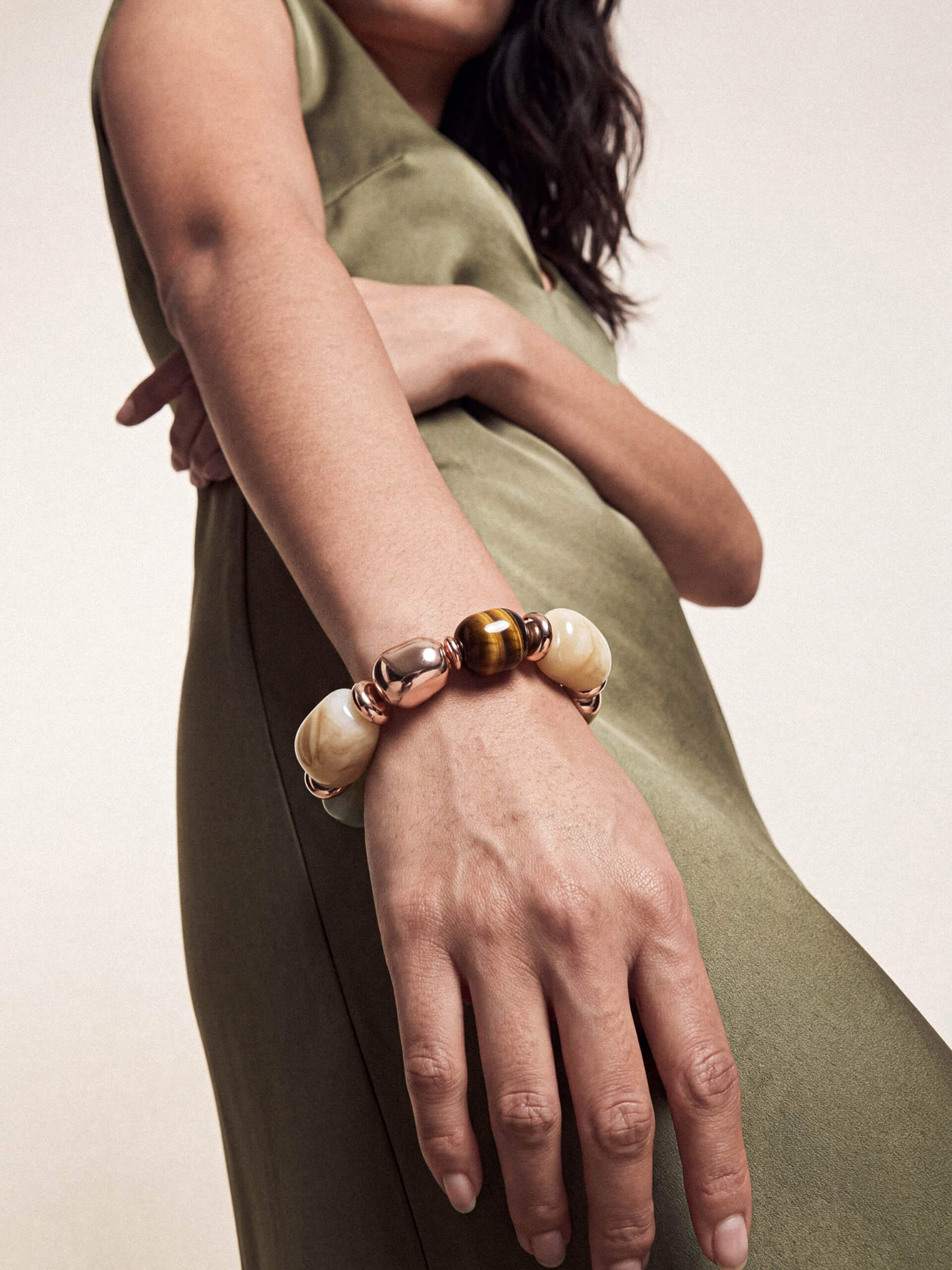 A close-up of a woman's wrist wearing a beaded bracelet with cream-colored round beads, a metallic rose gold bead, and a tiger's eye accent bead, set against an olive green dress.