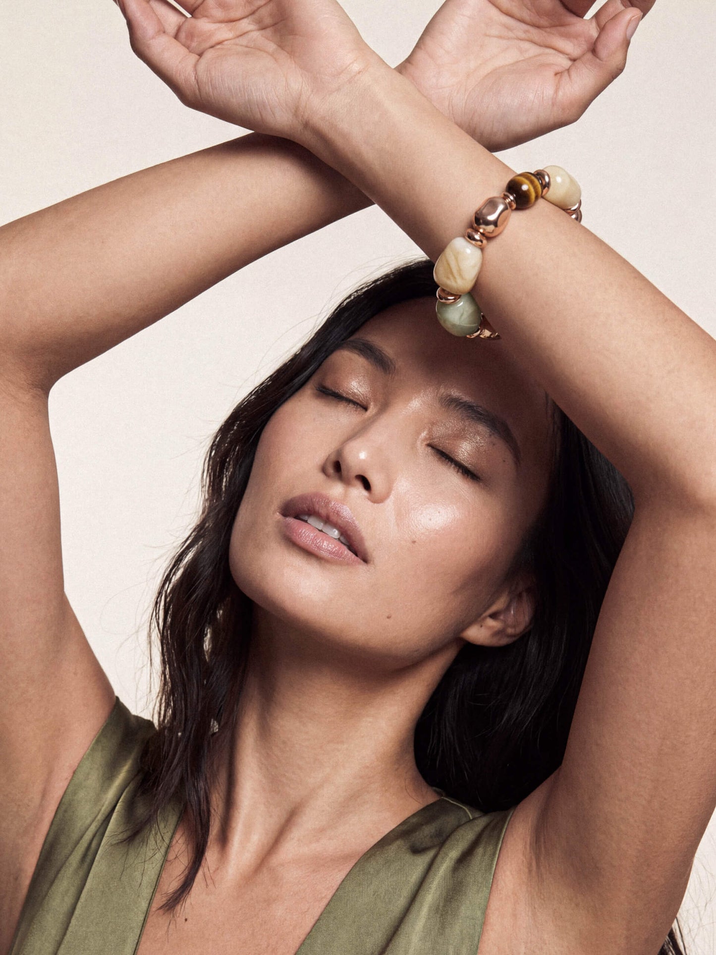 A woman with her arms crossed above her head wears a beaded bracelet with cream, green, and gold-toned beads on her wrist, set against a plain beige background.