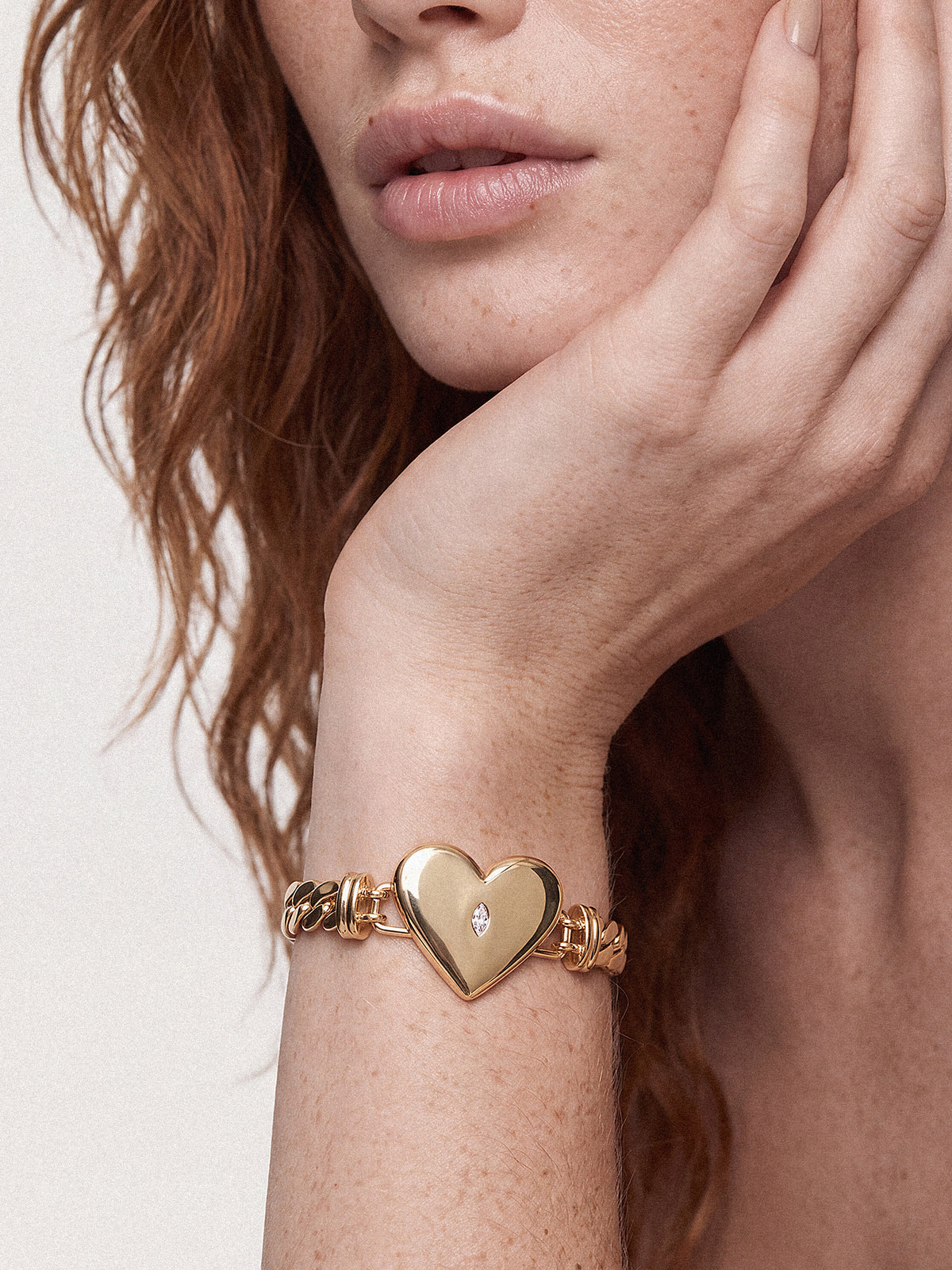 Close-up of a woman touching her face, wearing a gold chain bracelet with a heart-shaped charm and tiny diamond; intimate, elegant mood.