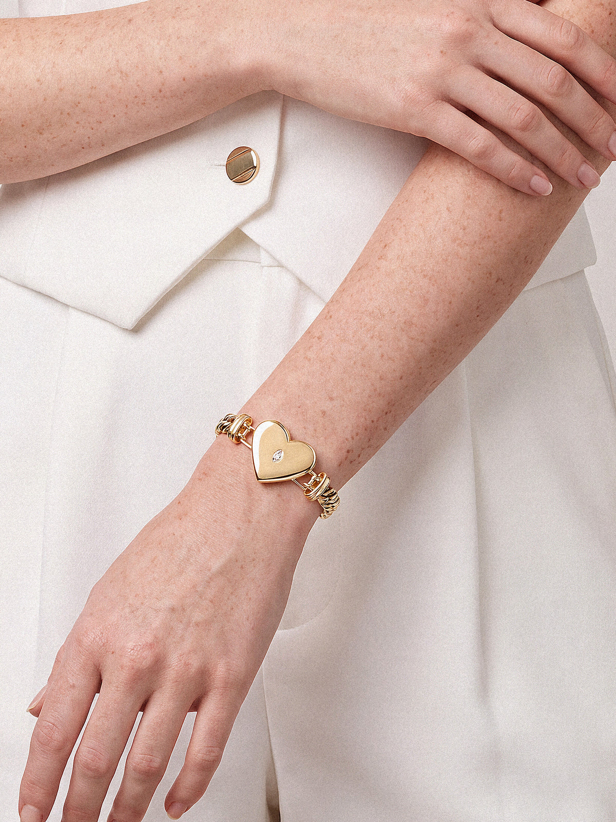 Close-up of crossed arms in a white outfit, featuring a gold bracelet with a heart-shaped charm; soft lighting creates an elegant tone.