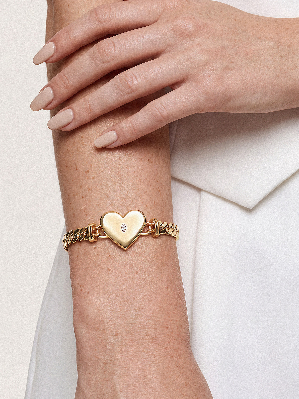 Close-up of crossed arms in a white outfit, featuring a gold bracelet with a heart-shaped charm; soft lighting creates an elegant tone.