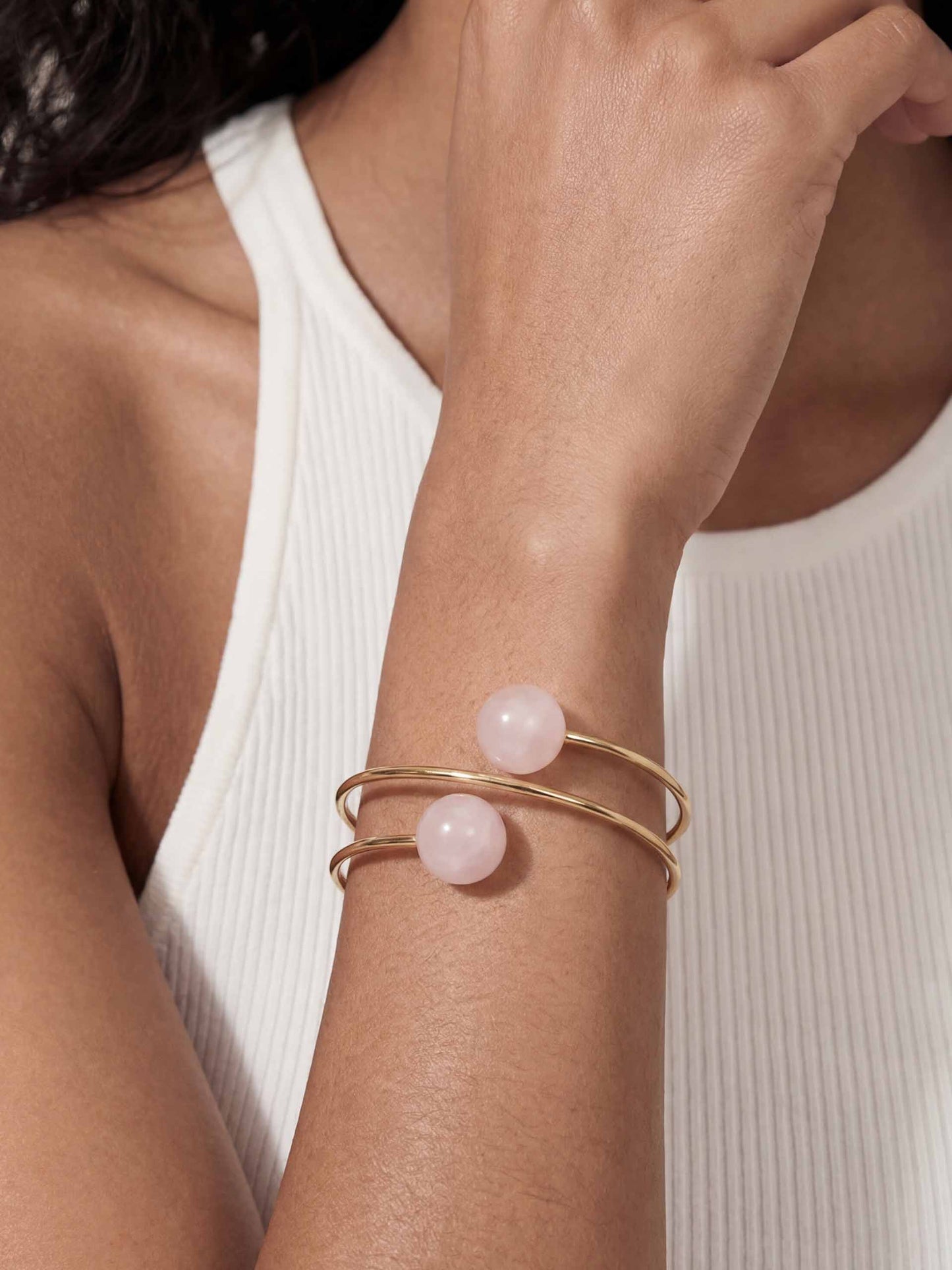 A person wearing a sleeveless white ribbed top showcases a gold bracelet with two pink beads. The tone is elegant and minimalistic.