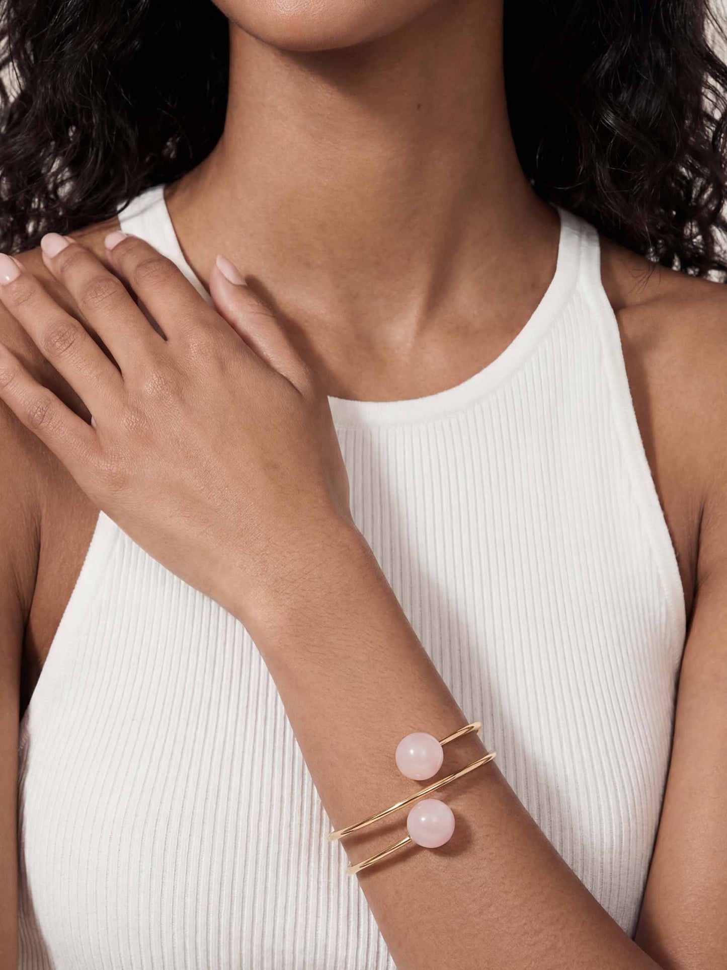 A person wearing a sleeveless white ribbed top showcases a gold bracelet with two pink beads. The tone is elegant and minimalistic.