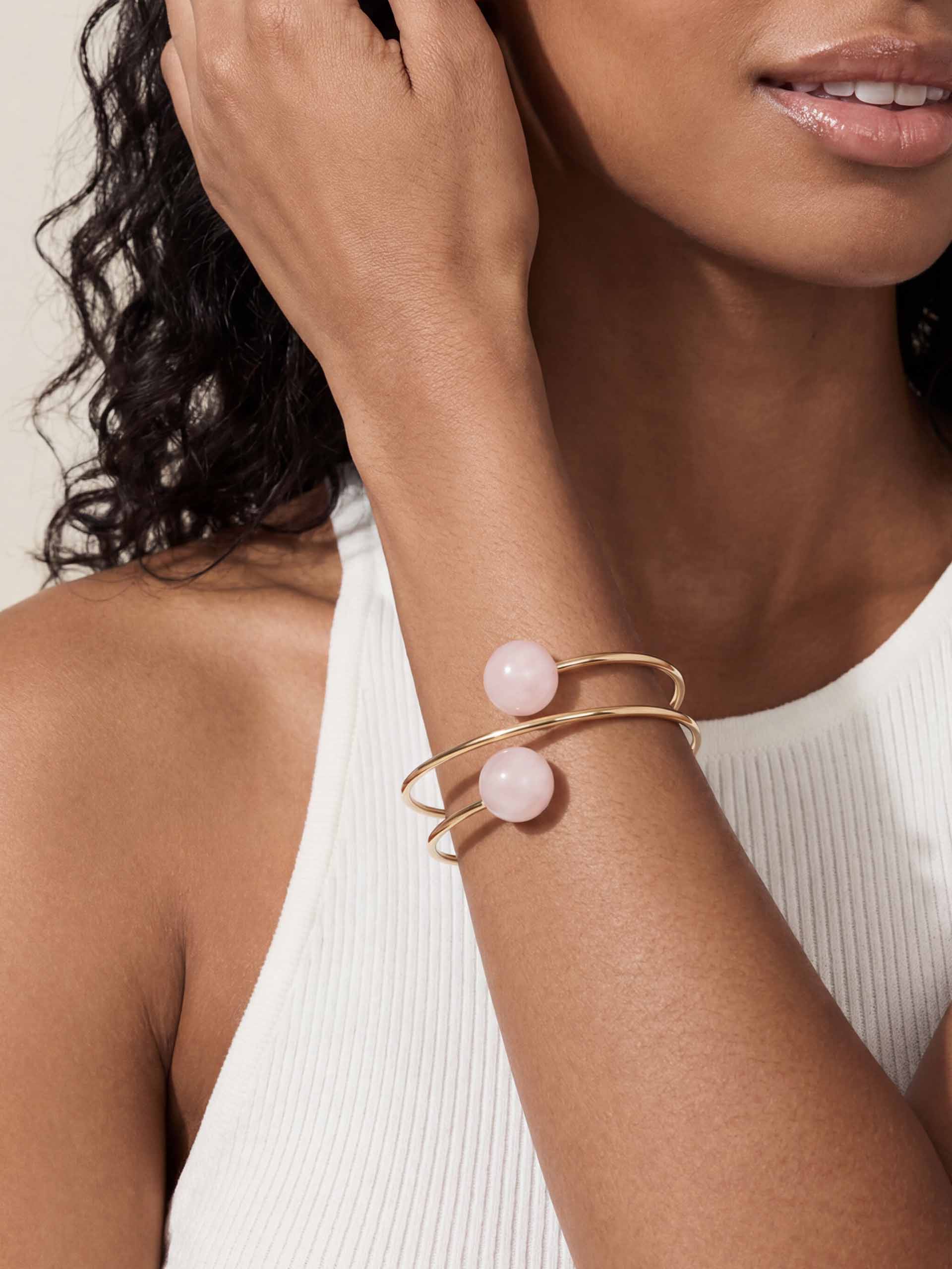 A person wearing a sleeveless white ribbed top showcases a gold bracelet with two pink beads. The tone is elegant and minimalistic.