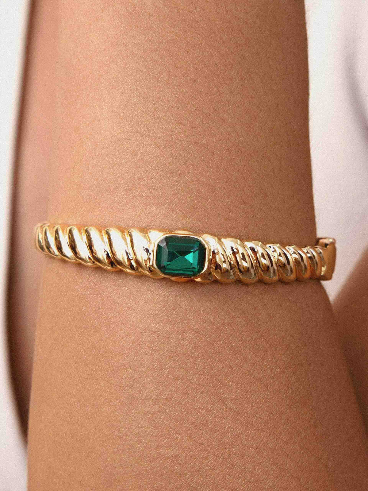 Close-up of a gold twisted bangle bracelet with a square green gemstone, worn on a wrist; warm lighting highlights its elegant design.