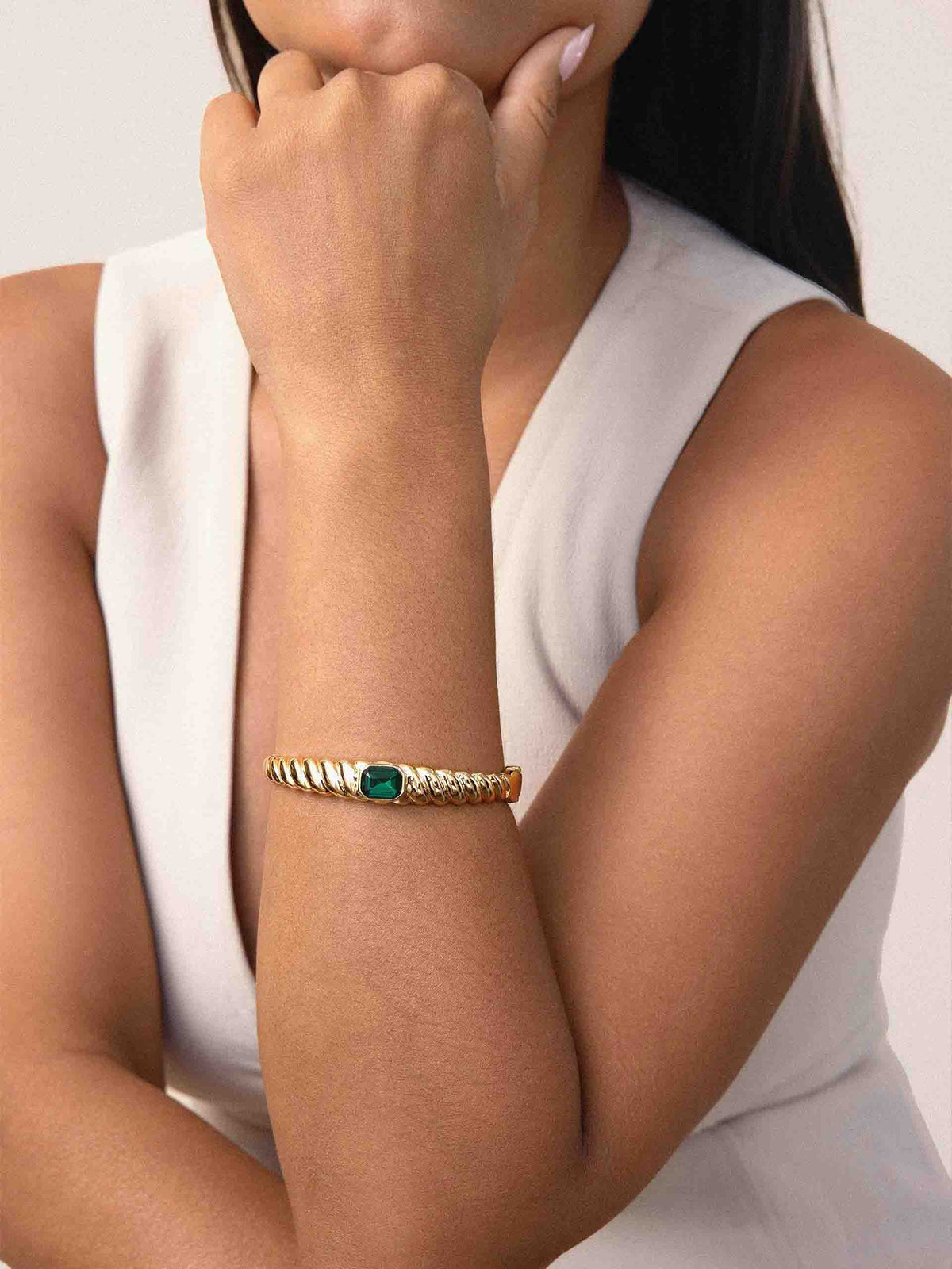 Close-up of a woman in a white sleeveless top wearing a gold bracelet with a green gemstone, posed thoughtfully with her hand near her chin.
