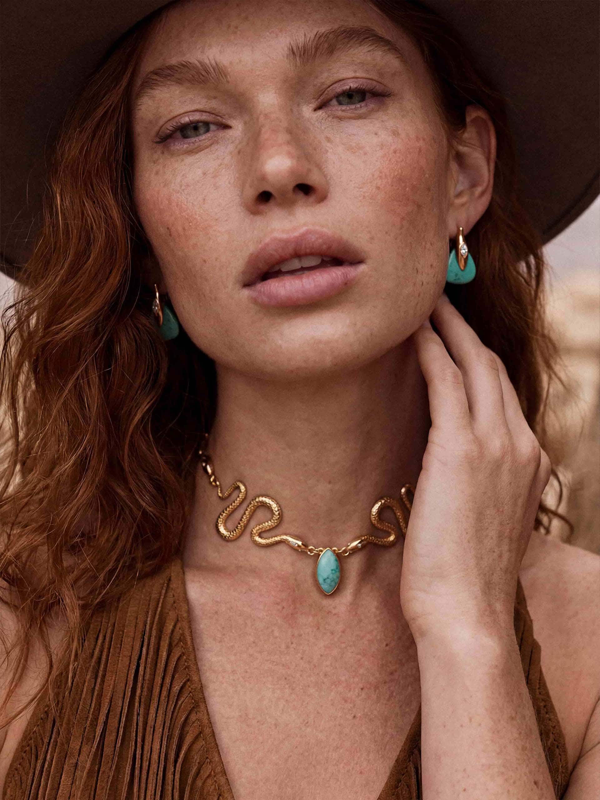 Portrait of woman in hat wearing turquoise earrings and gold snake necklace with desert backdrop