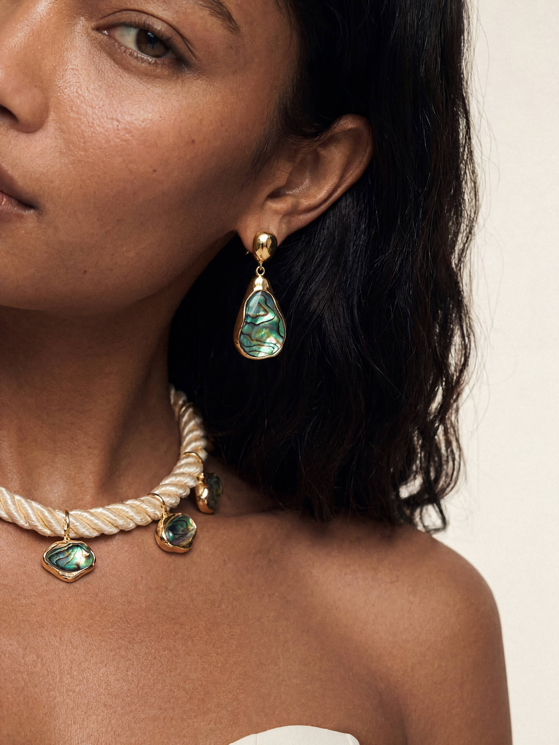 A model wearing a teardrop-shaped abalone earring with gold accents and matching chunky rope necklace with abalone pendants