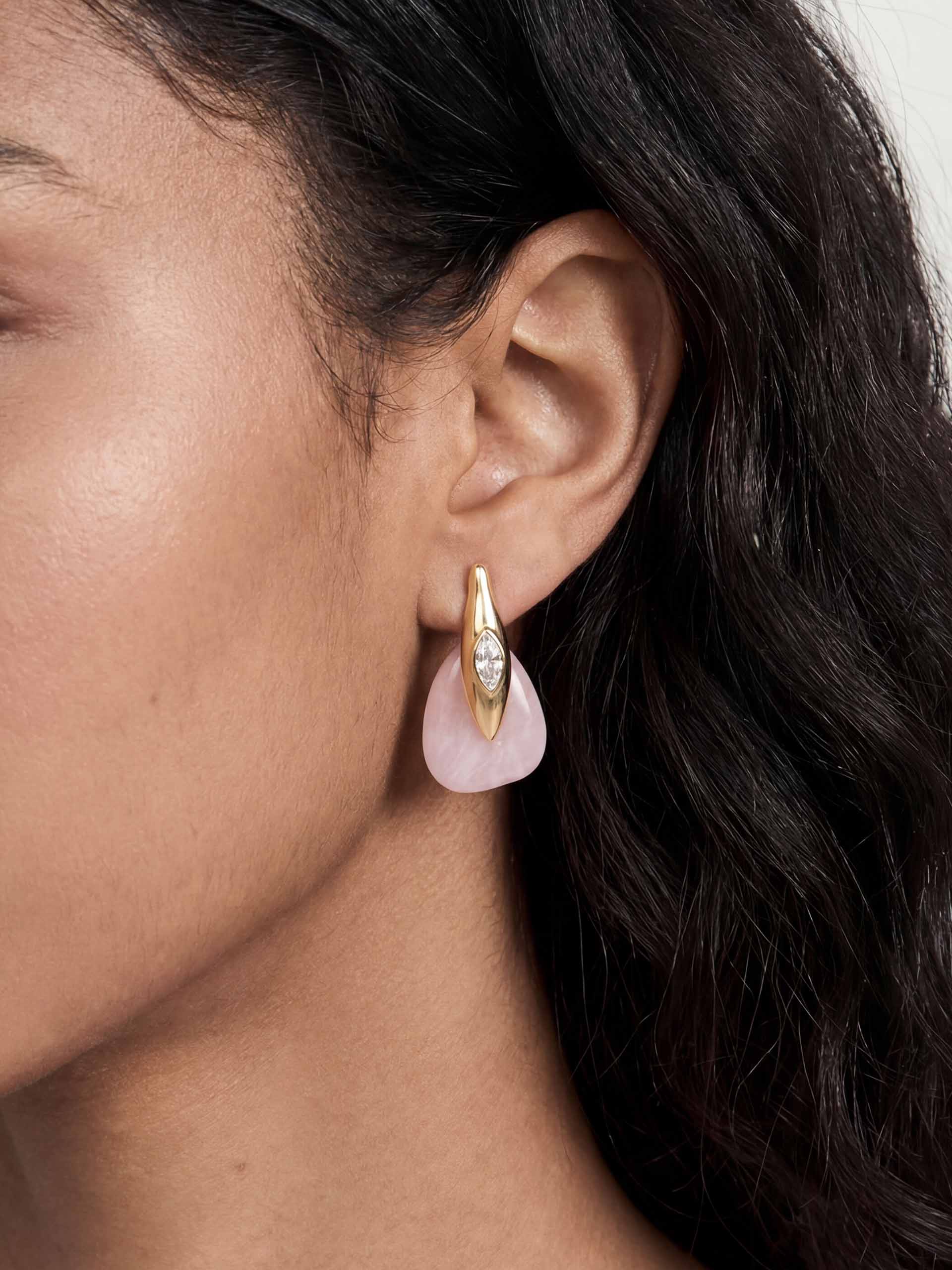 Close-up of a gold teardrop earring with a gemstone, featuring a pink stone dangling below. It is worn by a person with dark hair, suggesting elegance.