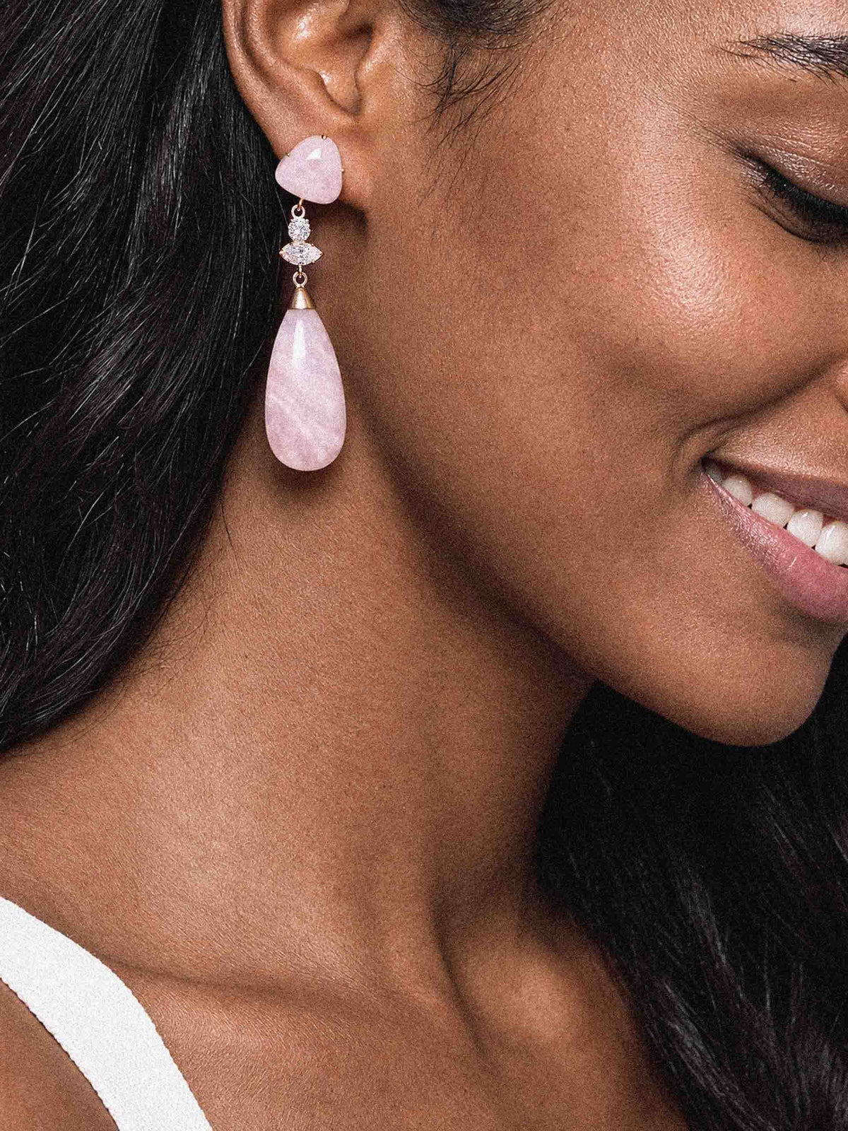 Close-up of a smiling woman wearing a delicate pink gemstone drop earring with sparkling silver accents, showcasing its elegant design against dark hair.