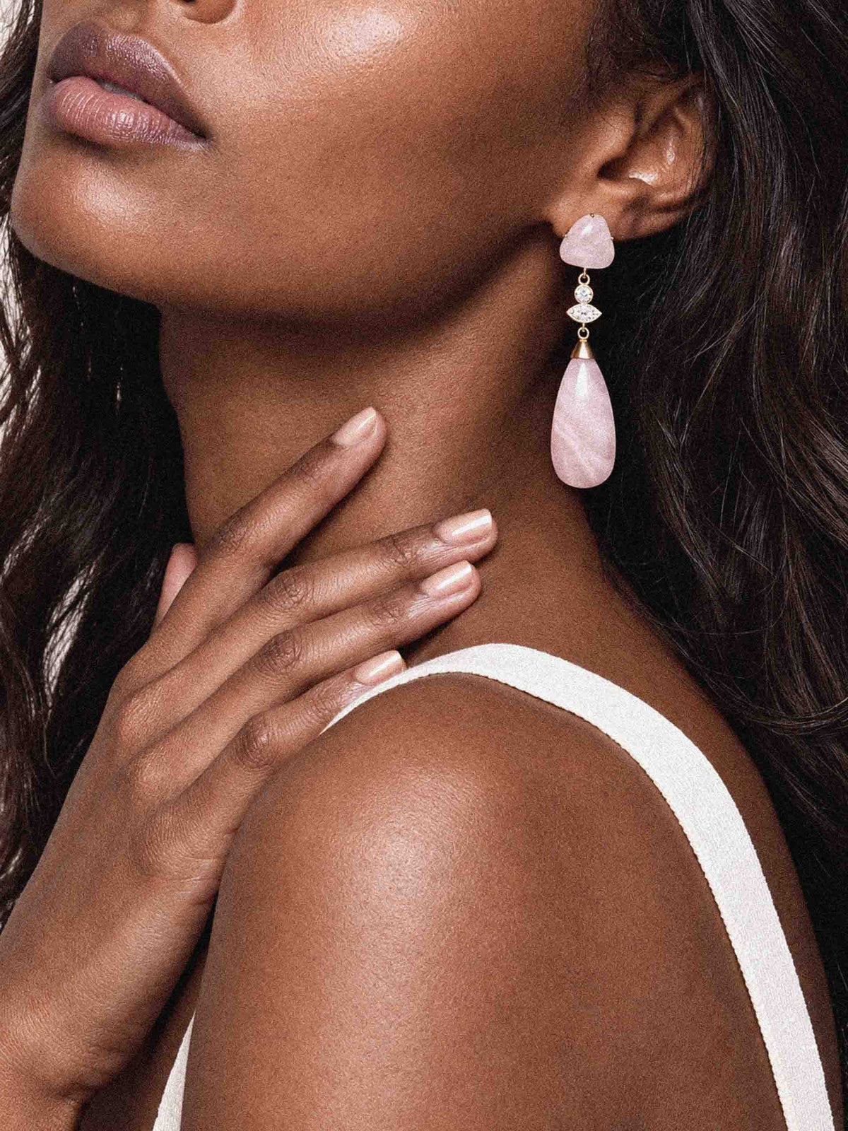 Close-up beauty portrait of a woman wearing elegant pink gemstone drop earrings, her hand resting on her neck; soft lighting creates a calm, refined mood.