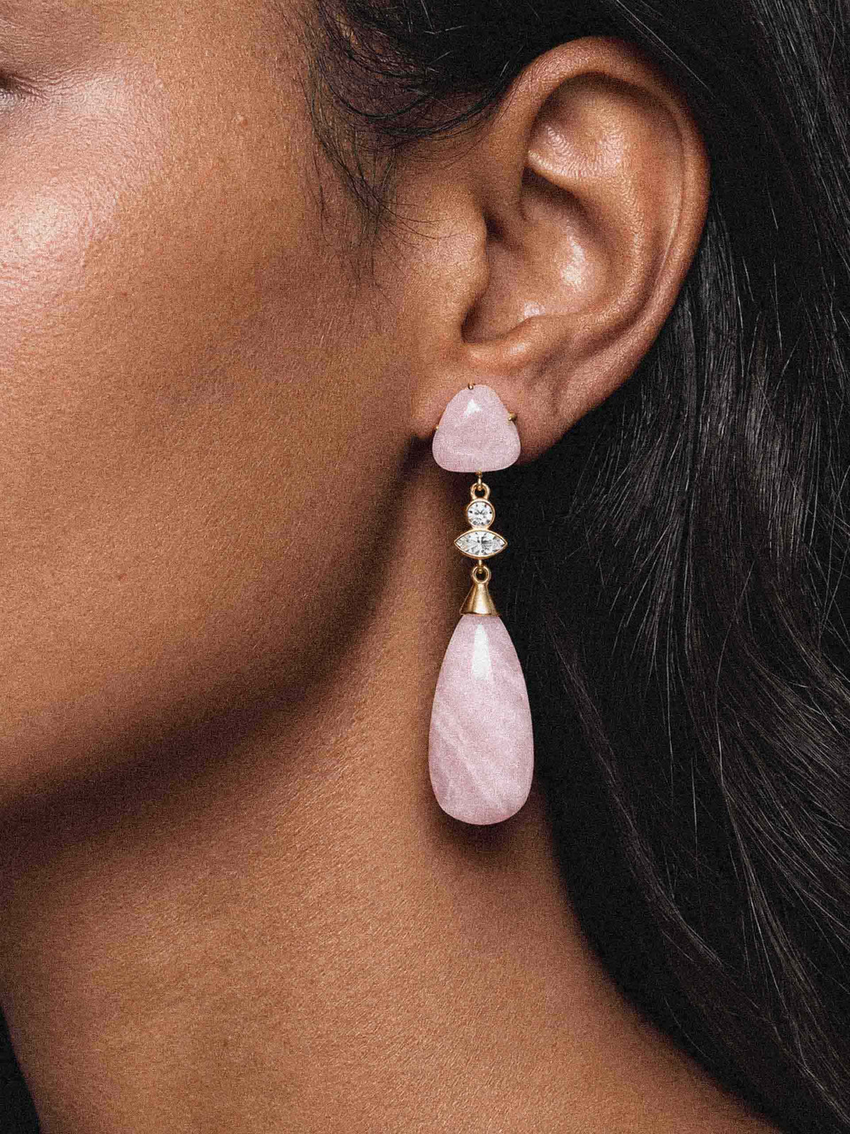 Close-up of a woman wearing elegant gold drop earrings with pale pink gemstones, set against dark hair; the jewelry looks delicate and refined.