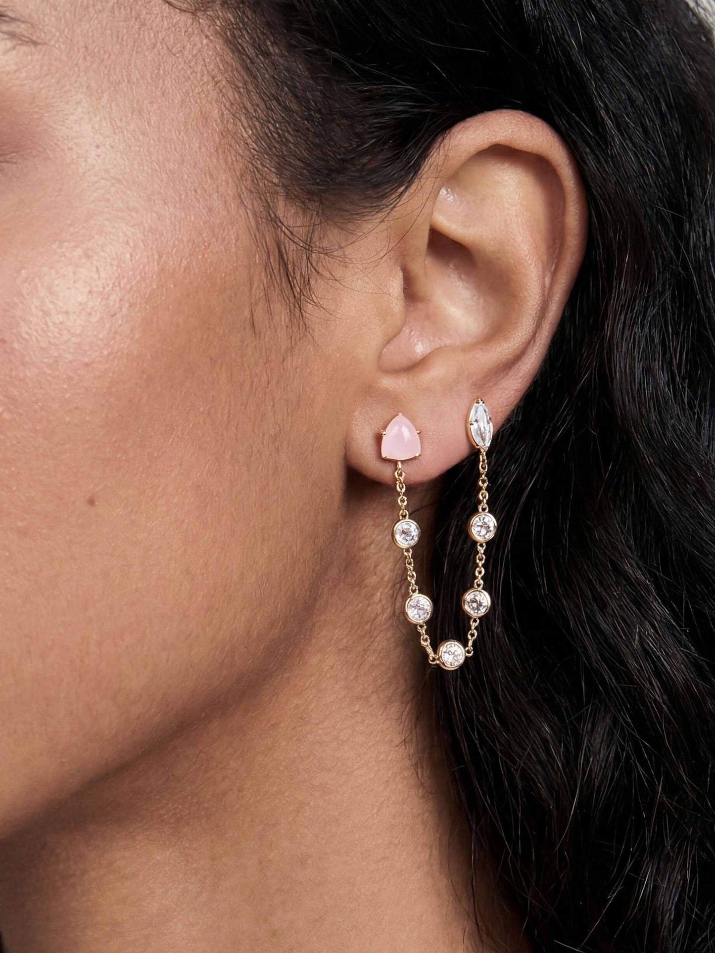 Close-up of a woman's ear wearing elegant gold earrings. One stud features a pink stone, and a chain with pearls loops beneath, conveying sophistication.