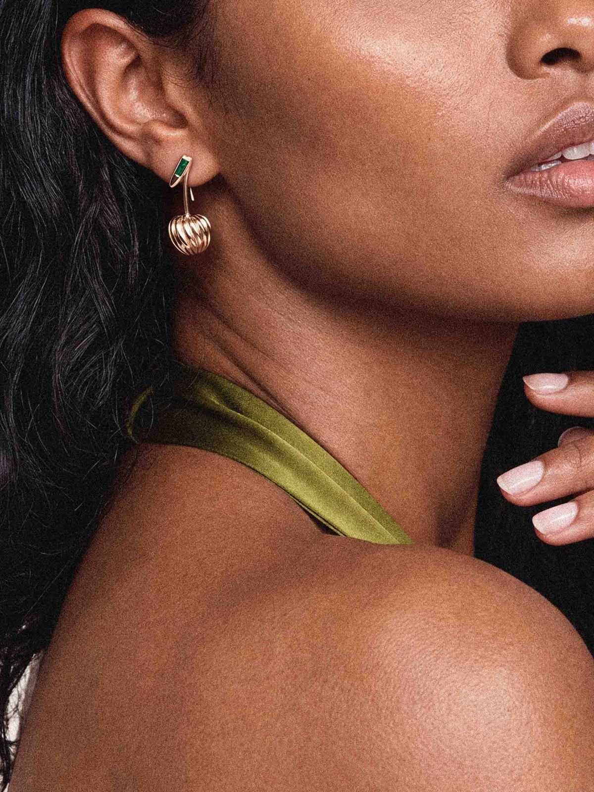 Close-up of a woman wearing a gold drop earring with a green accent, an olive-green strap, and pale pink nails; the portrait feels elegant and refined.