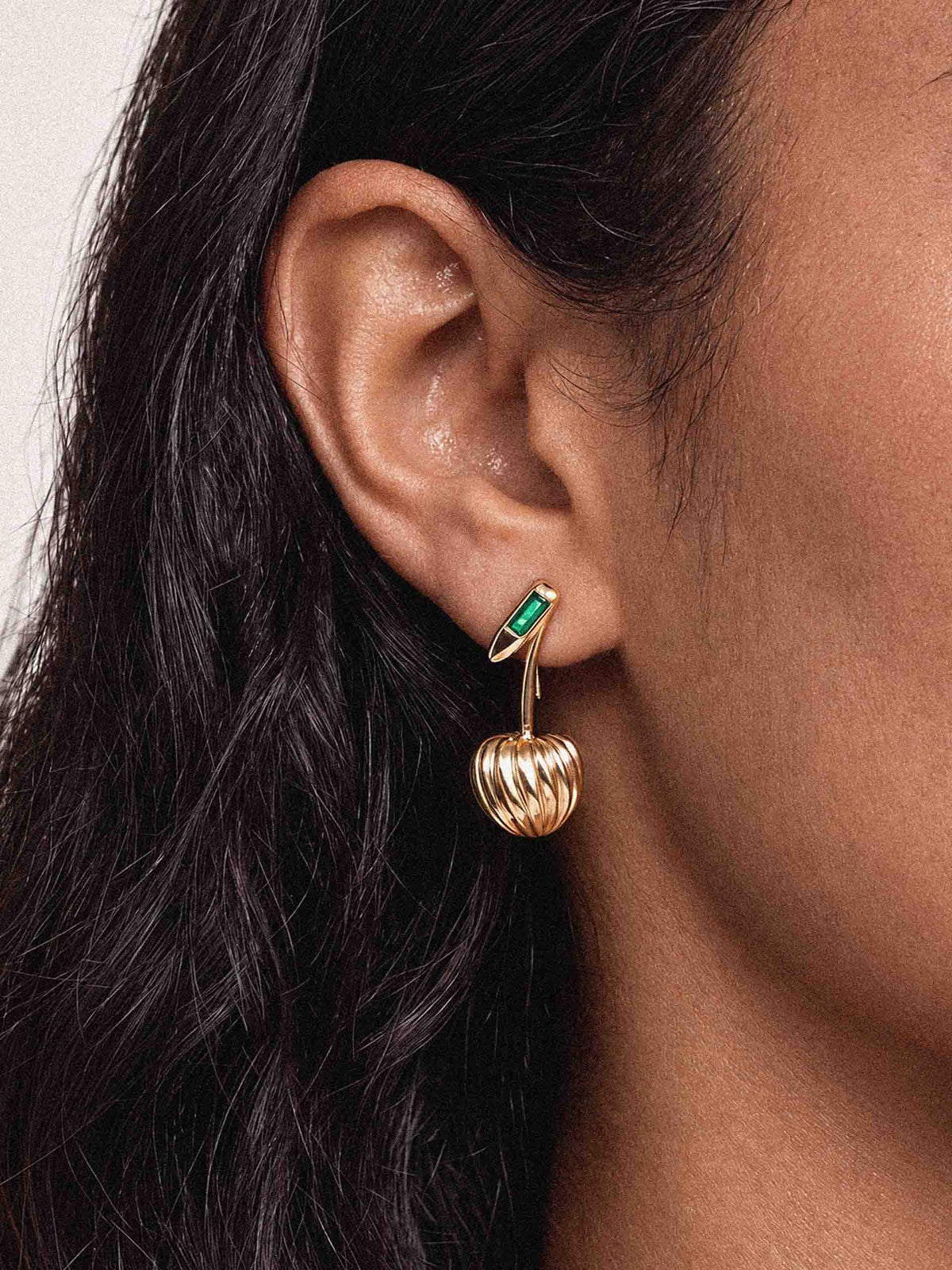 Close-up of a woman’s ear with long dark hair, wearing a delicate gold drop earring with a green accent and ribbed heart-shaped charm; elegant, refined mood.