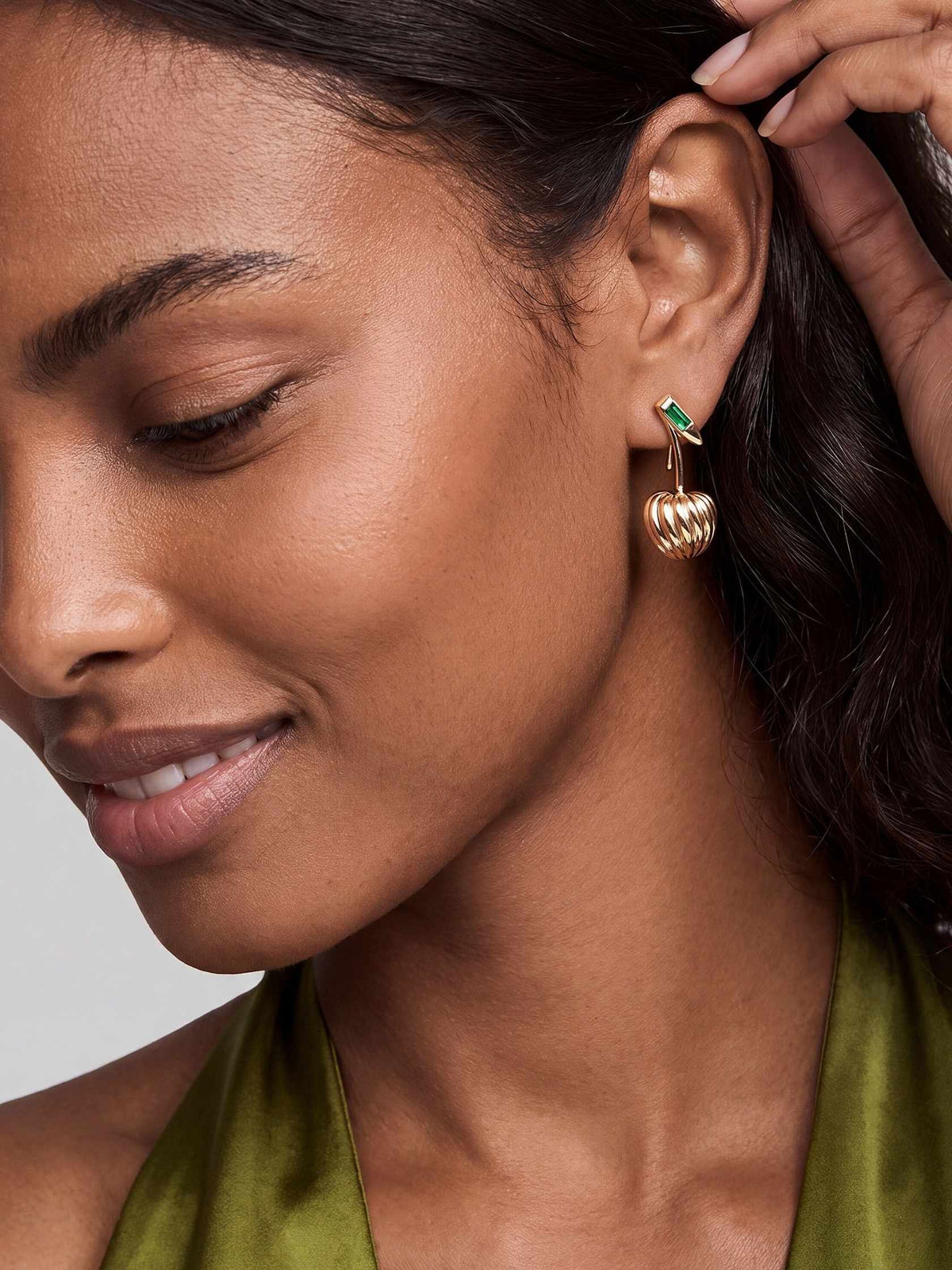 Close-up of a smiling woman with dark hair wearing a gold drop earring with a green stone, showing its sculpted design against her skin.