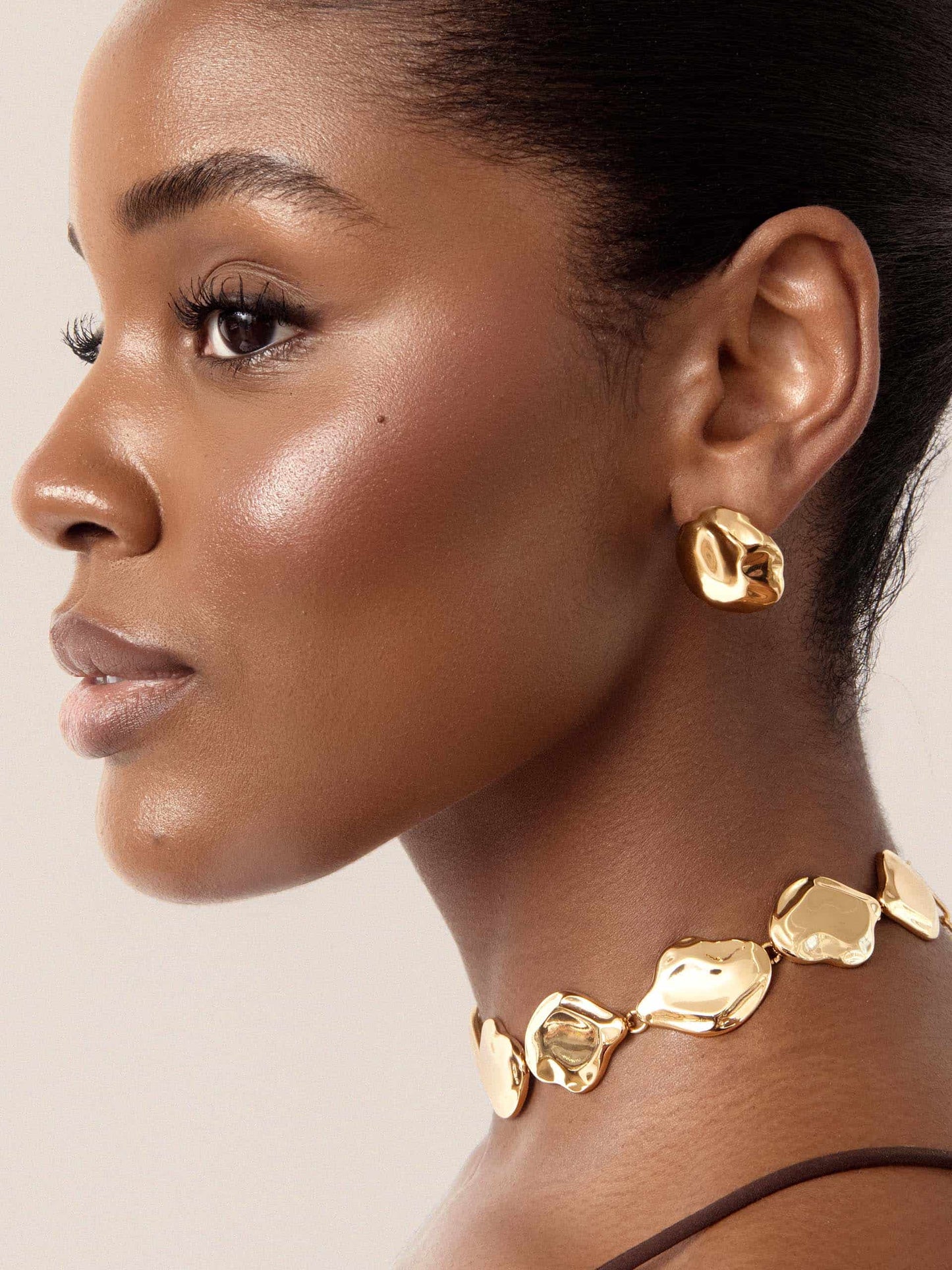 Close-up of a model wearing gold earrings and a gold necklace against a neutral background 2