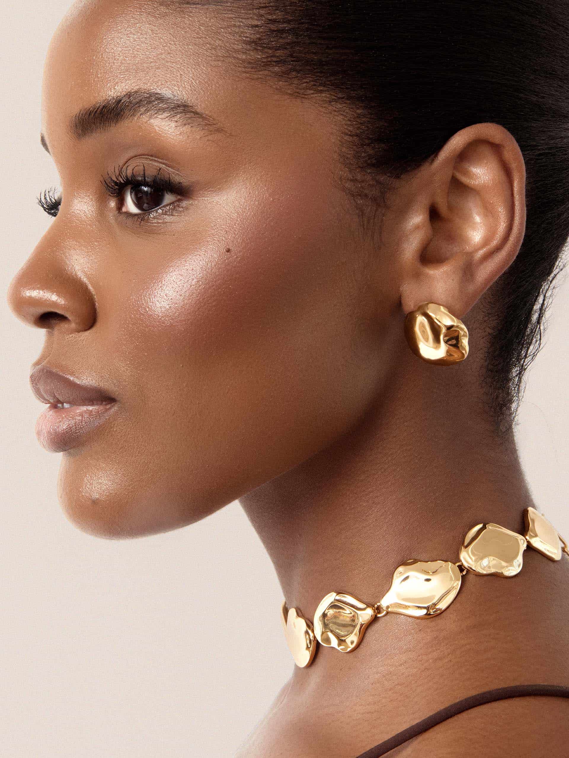 Close-up of a model wearing gold earrings and a gold necklace against a neutral background 2