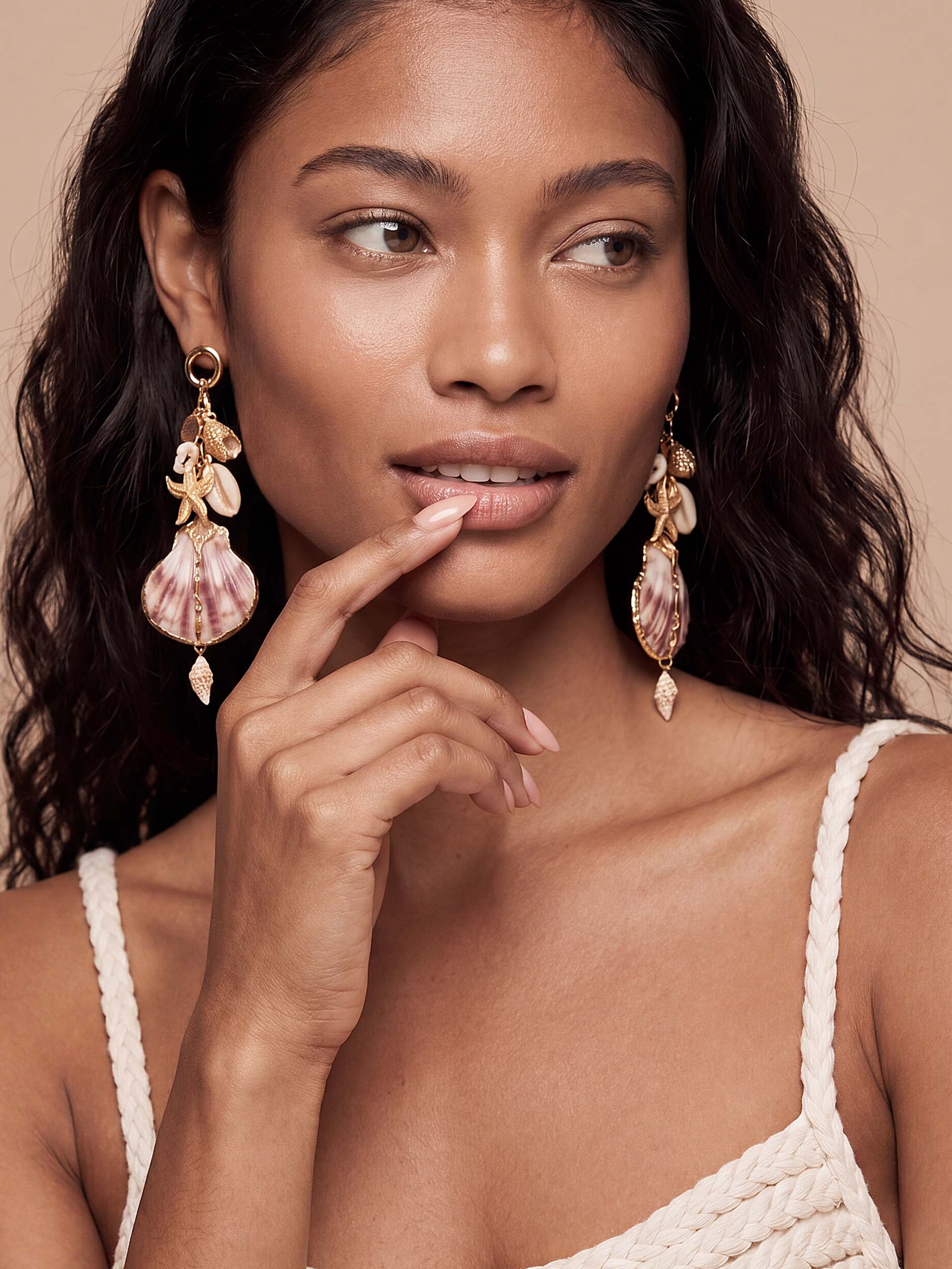 Woman with wavy hair wearing a cream dress and large sahell earrings touching her lip with her finger