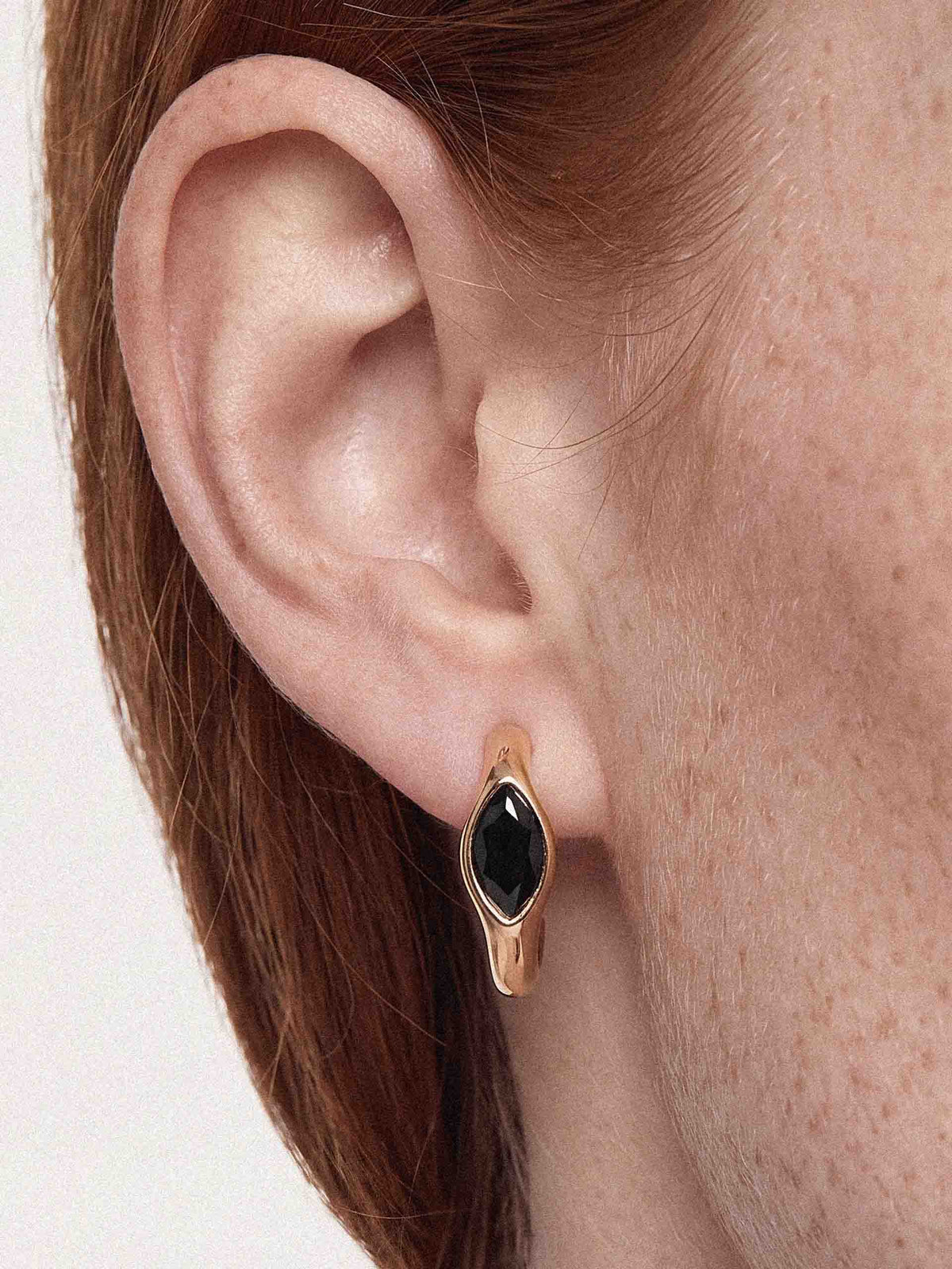 Close-up of a woman’s ear with brown hair and a black oval gold earring, highlighting detailed skin texture and elegant jewelry.
