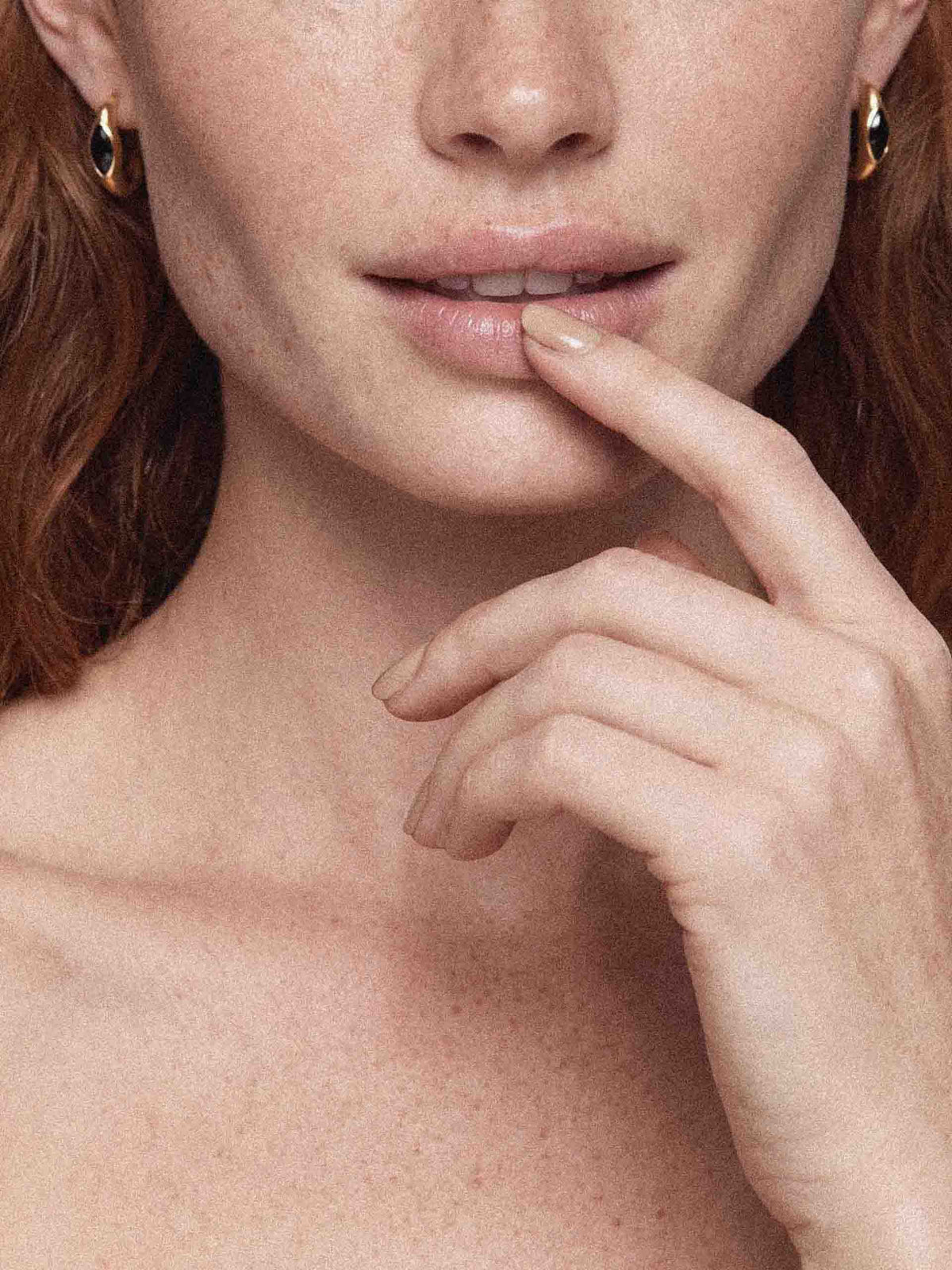Close-up of a freckled woman touching her lips with one finger, framed by auburn hair and gold hoop earrings; warm, intimate beauty tone.