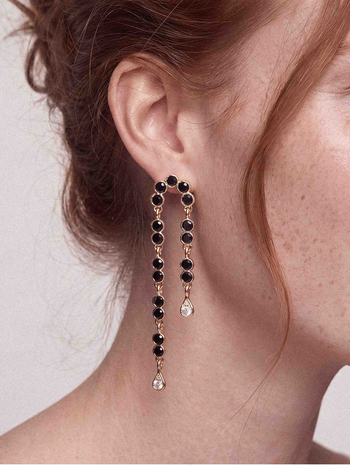 Close-up of a woman’s ear wearing elegant gold drop earrings with black gemstones and teardrop crystals, creating a refined, glamorous look.