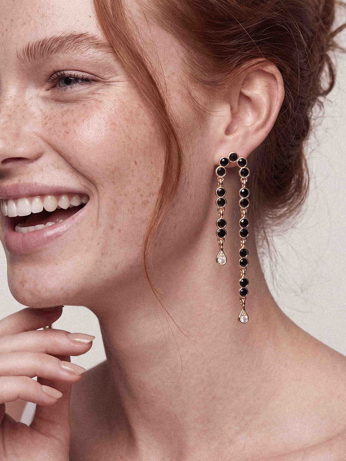 Close-up portrait of a smiling, freckled red-haired woman wearing long black-and-gold dangling earrings, hand near chin; warm, elegant mood.