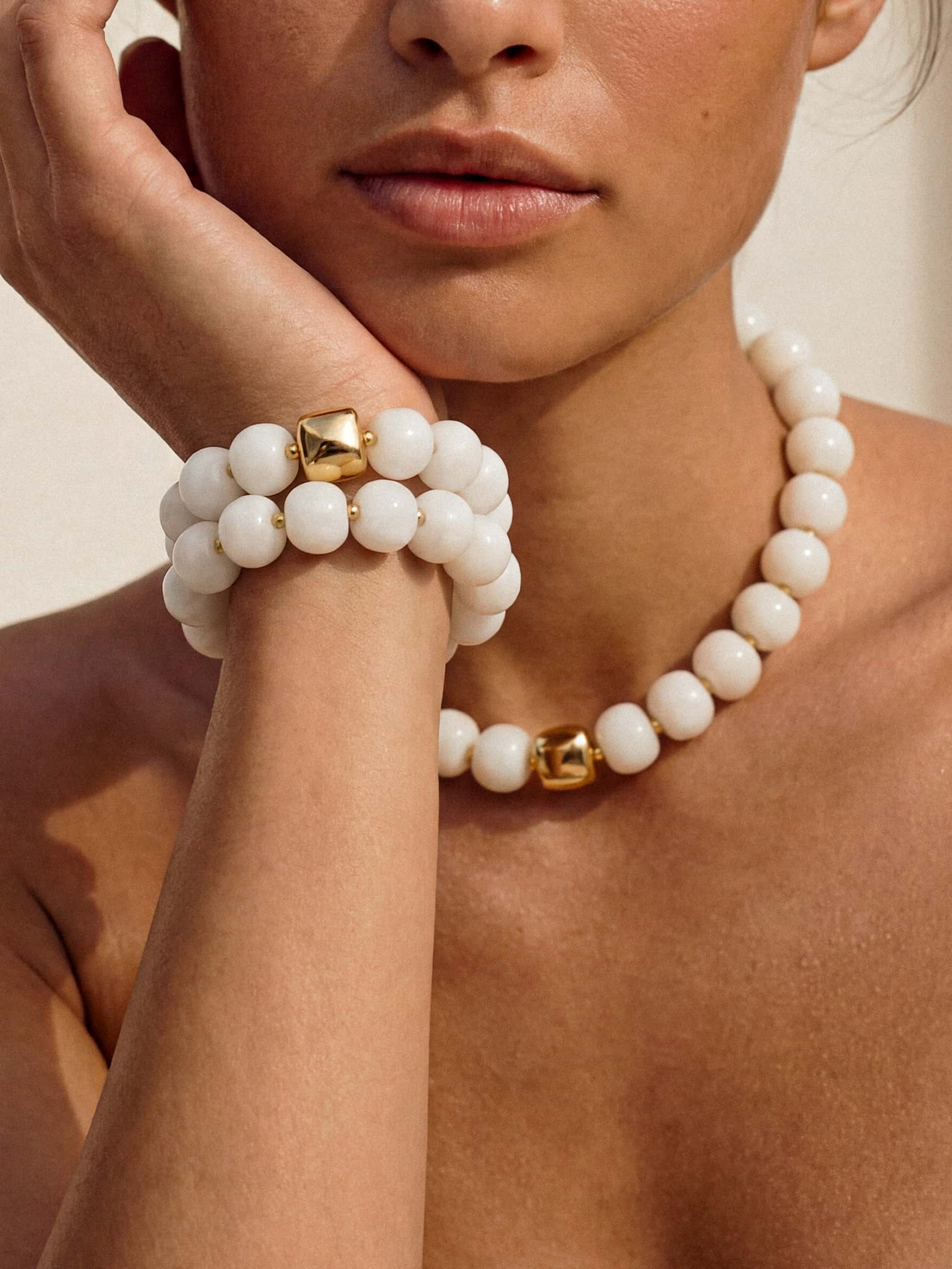 Model holding her face wearing a matching white bead necklace and bracelet.