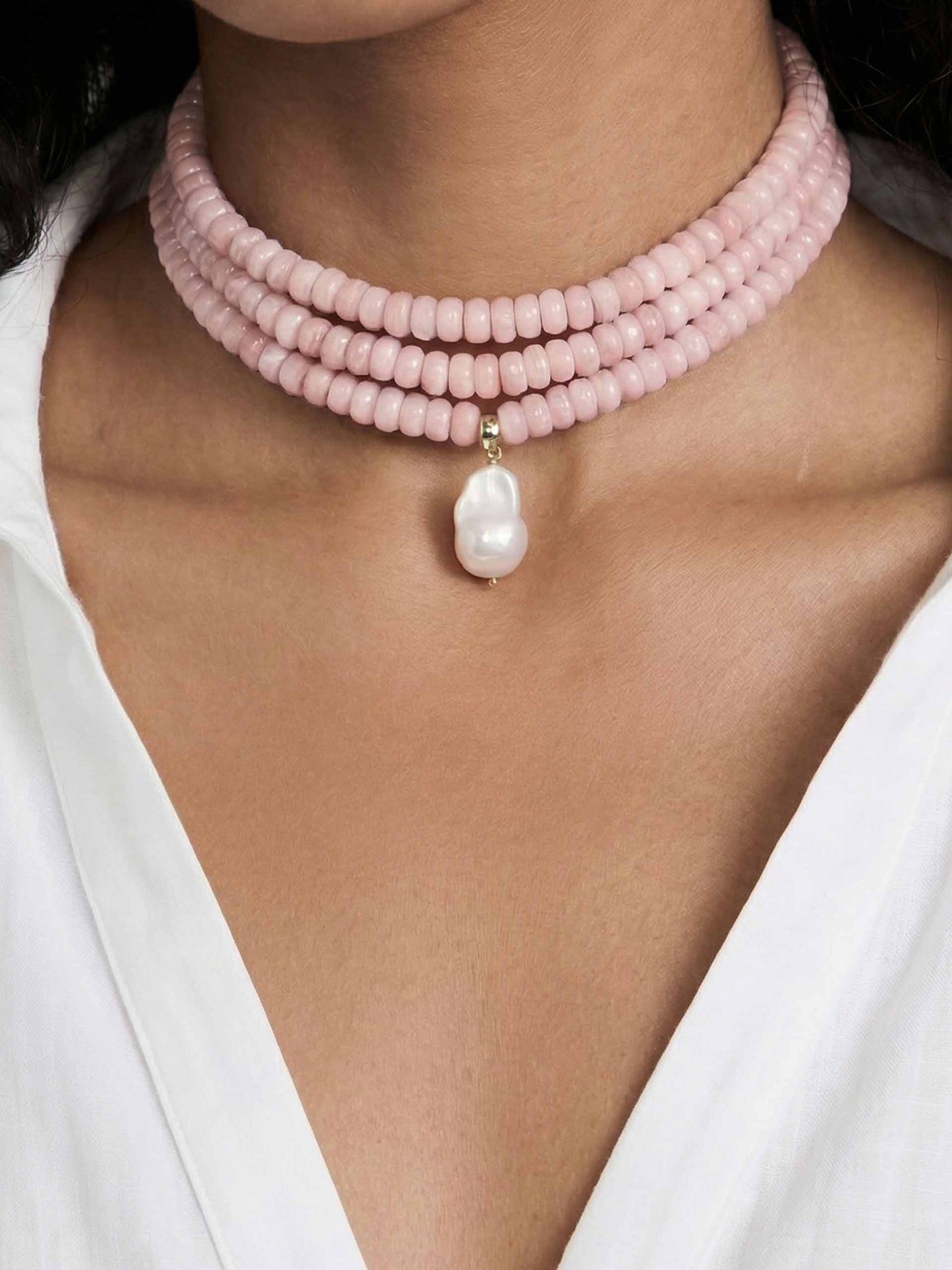 A woman wears a three-strand pink bead choker with a dangling white pearl pendant. She is dressed in a white blouse, creating an elegant, sophisticated look.