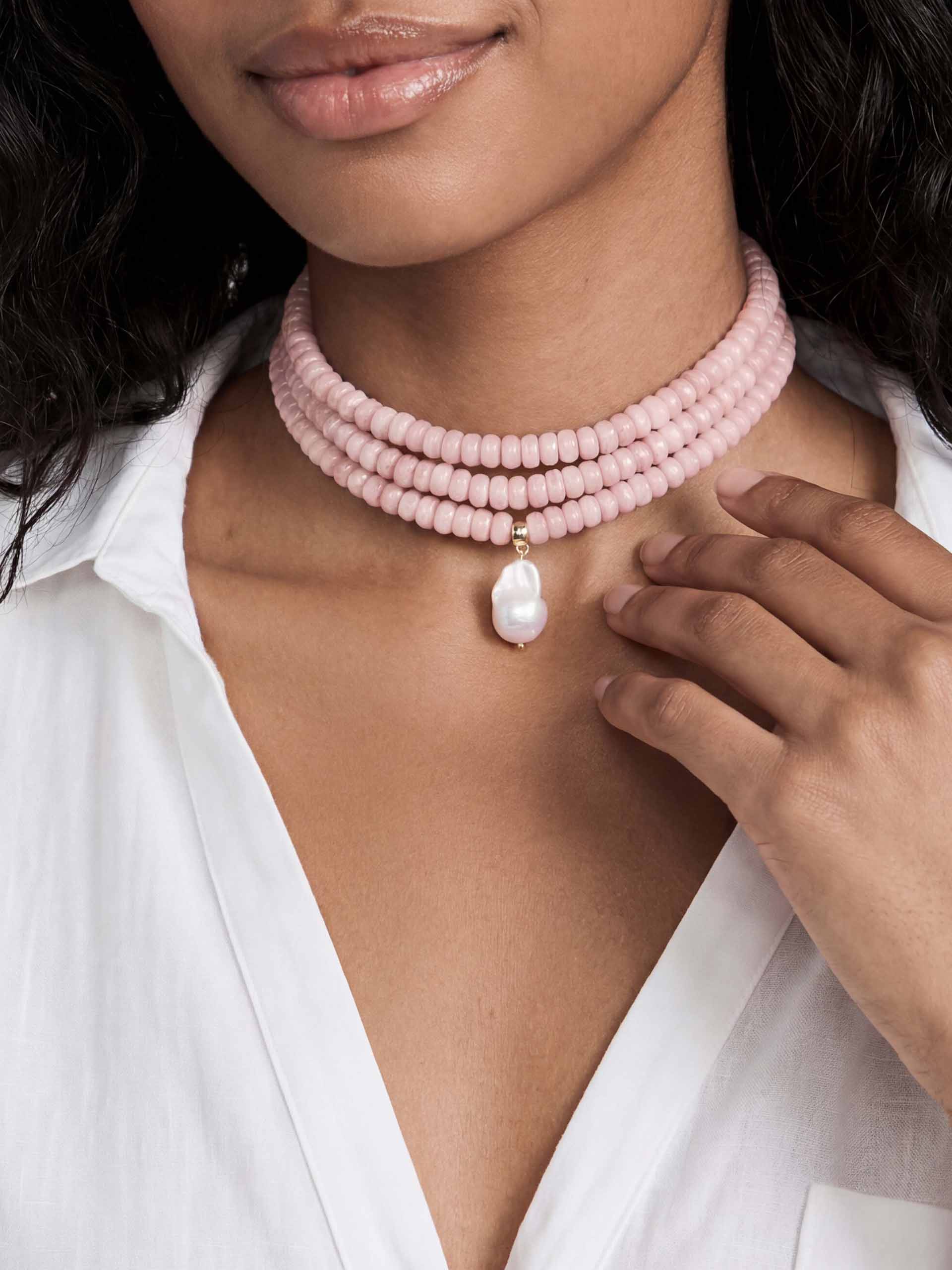 A woman wears a three-strand pink bead choker with a dangling white pearl pendant. She is dressed in a white blouse, creating an elegant, sophisticated look.