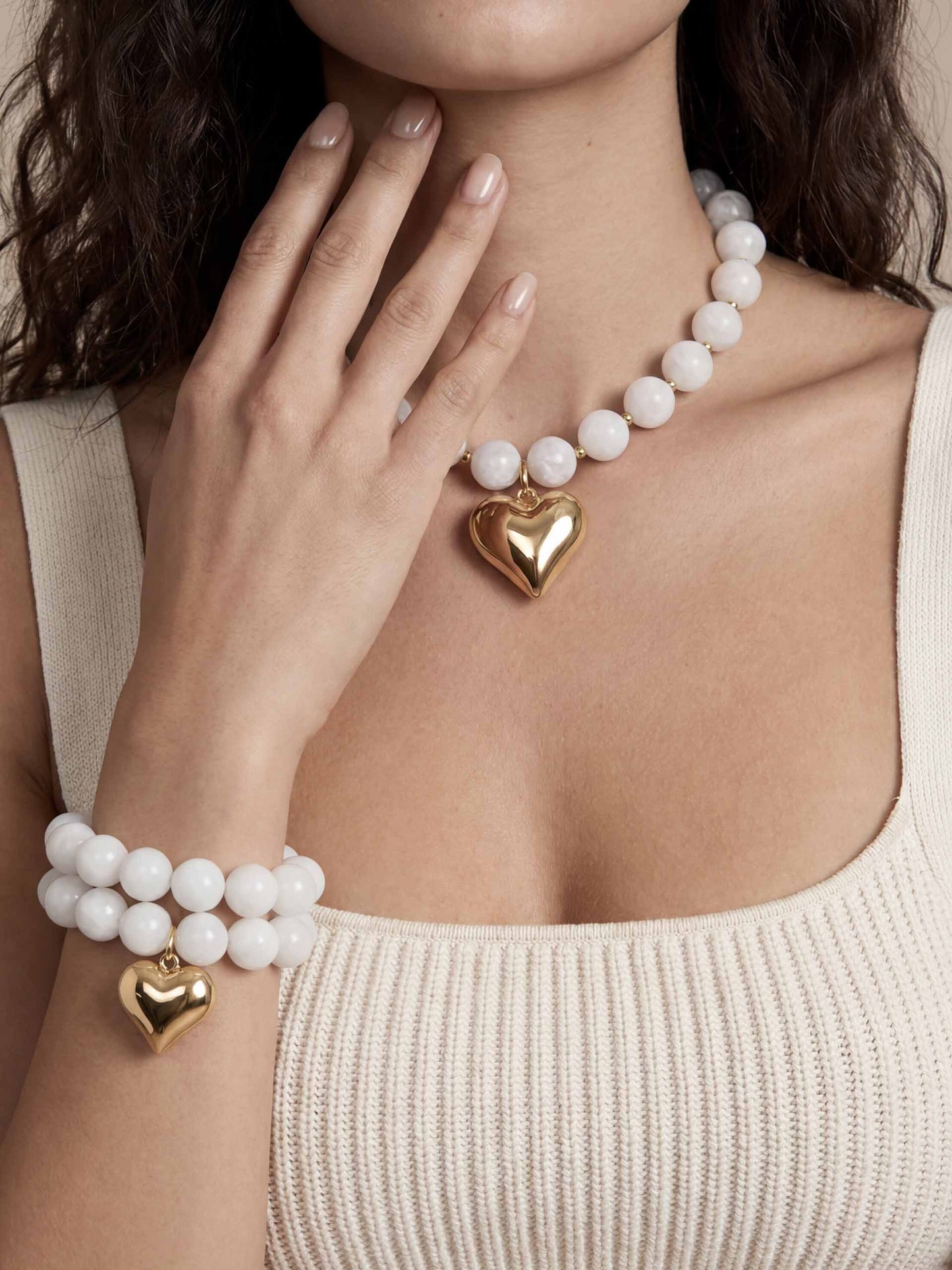 A woman wearing a cream tank top showcases a white bead bone necklace and bracelet, each with a gold heart charm. Her hand gently touches her neck, conveying elegance.