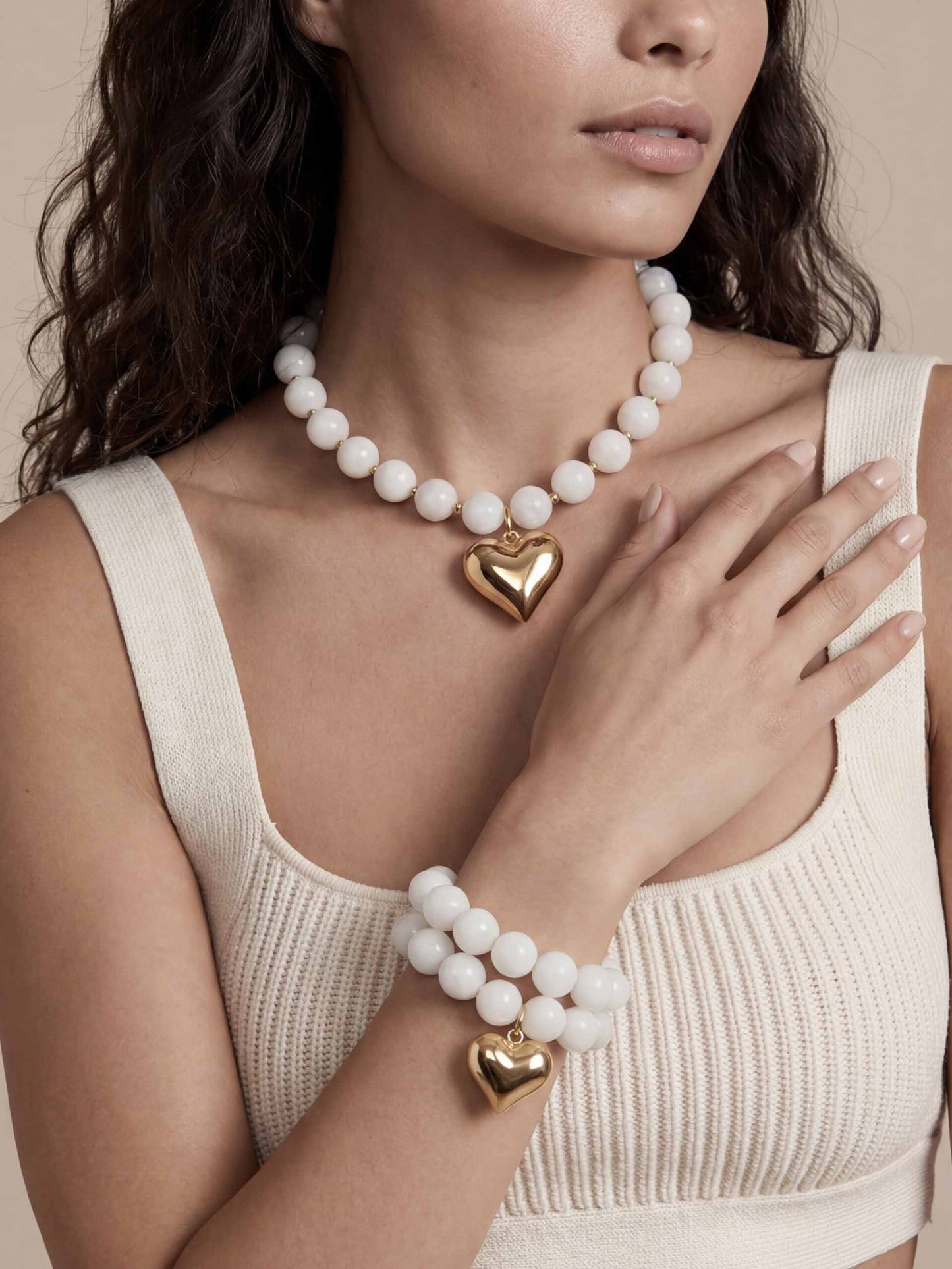 Woman wearing a cream knit top, accessorized with a pearl necklace and bracelet, both featuring gold heart pendants.