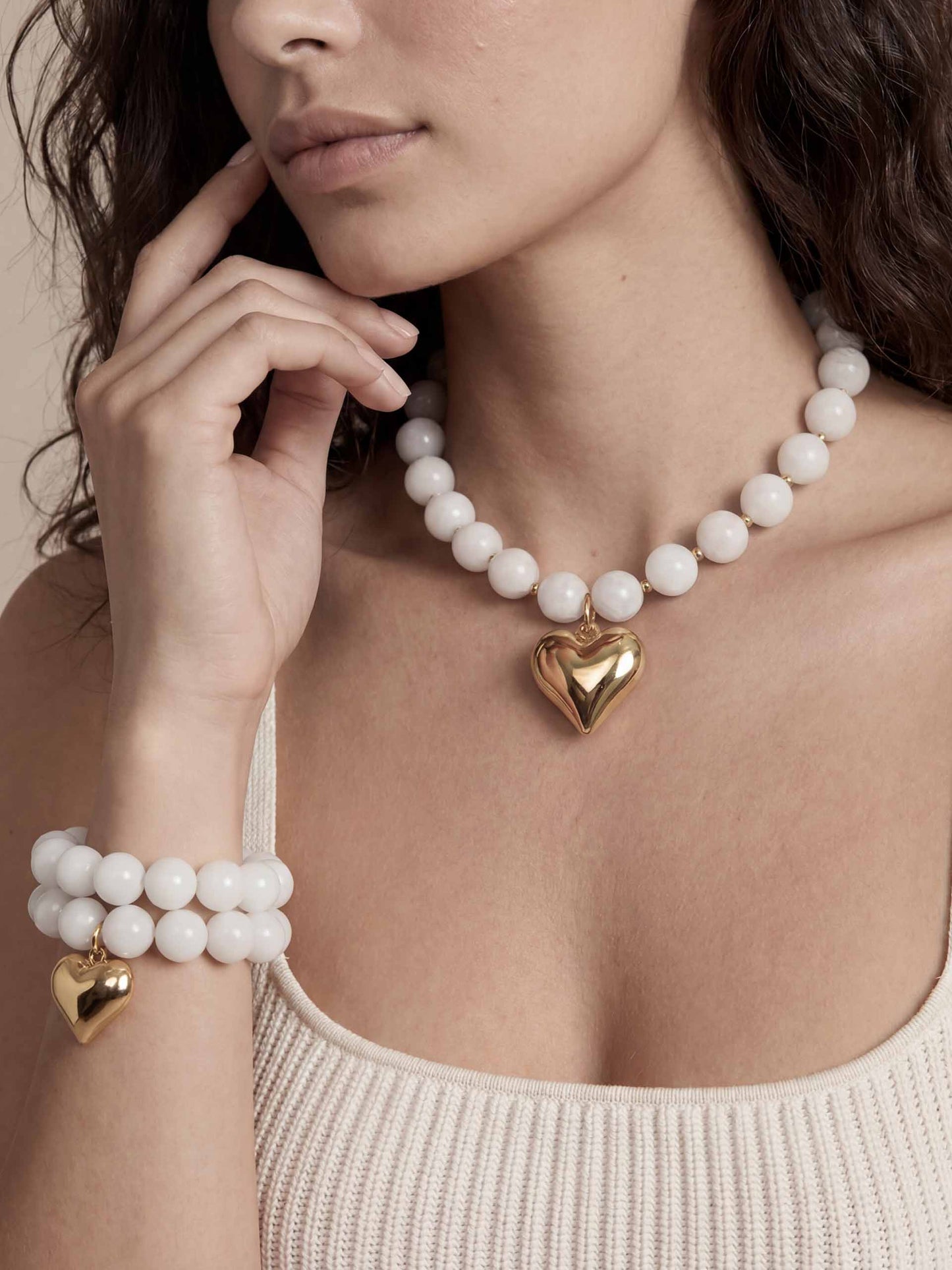 A woman wearing a beaded bone necklace and bracelet set with large white beads, each featuring a gold heart pendant. 
