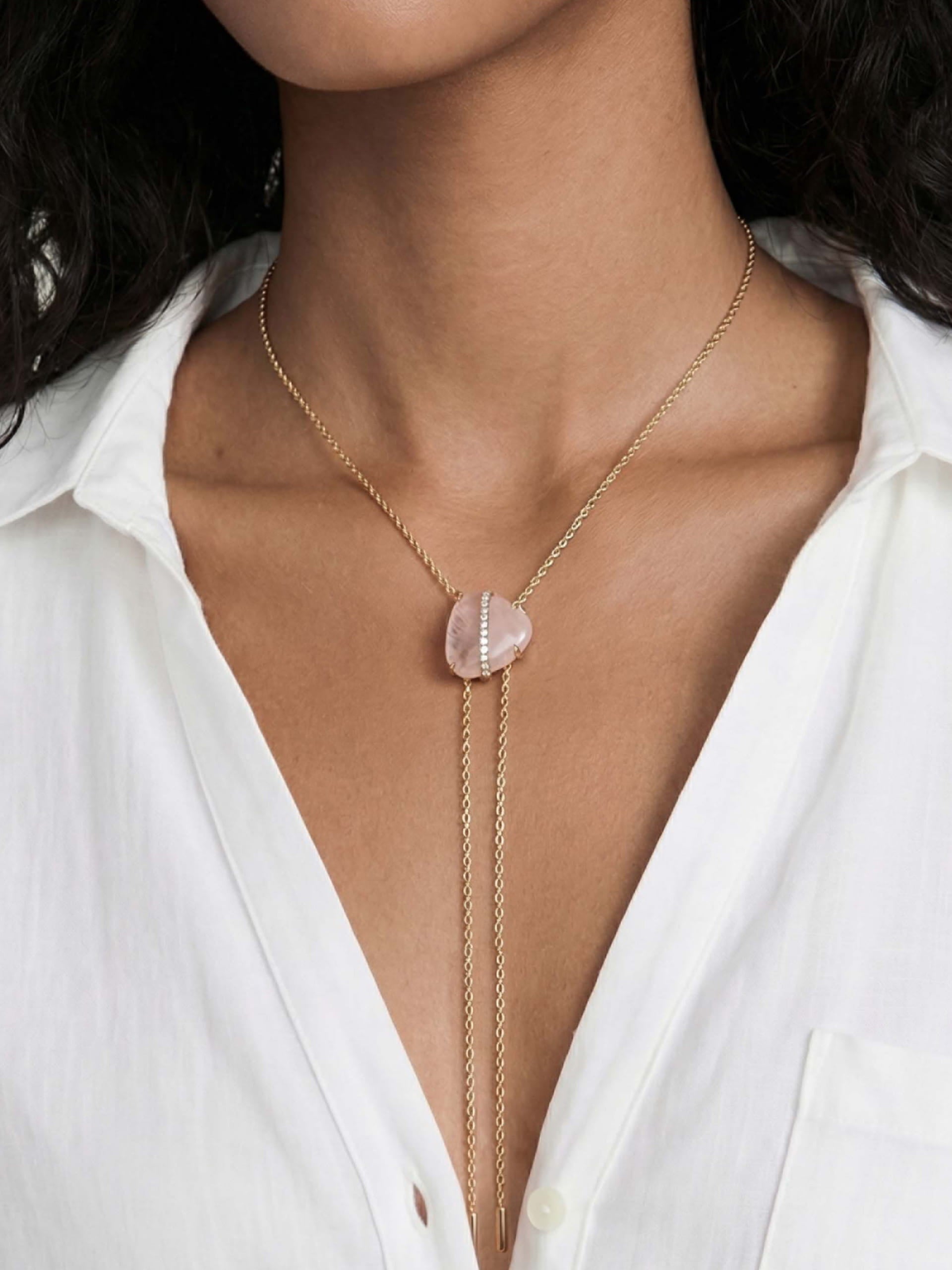 A woman wearing a white shirt displays a gold necklace with a pink stone pendant. The necklace features delicate chain tassels, creating an elegant look.