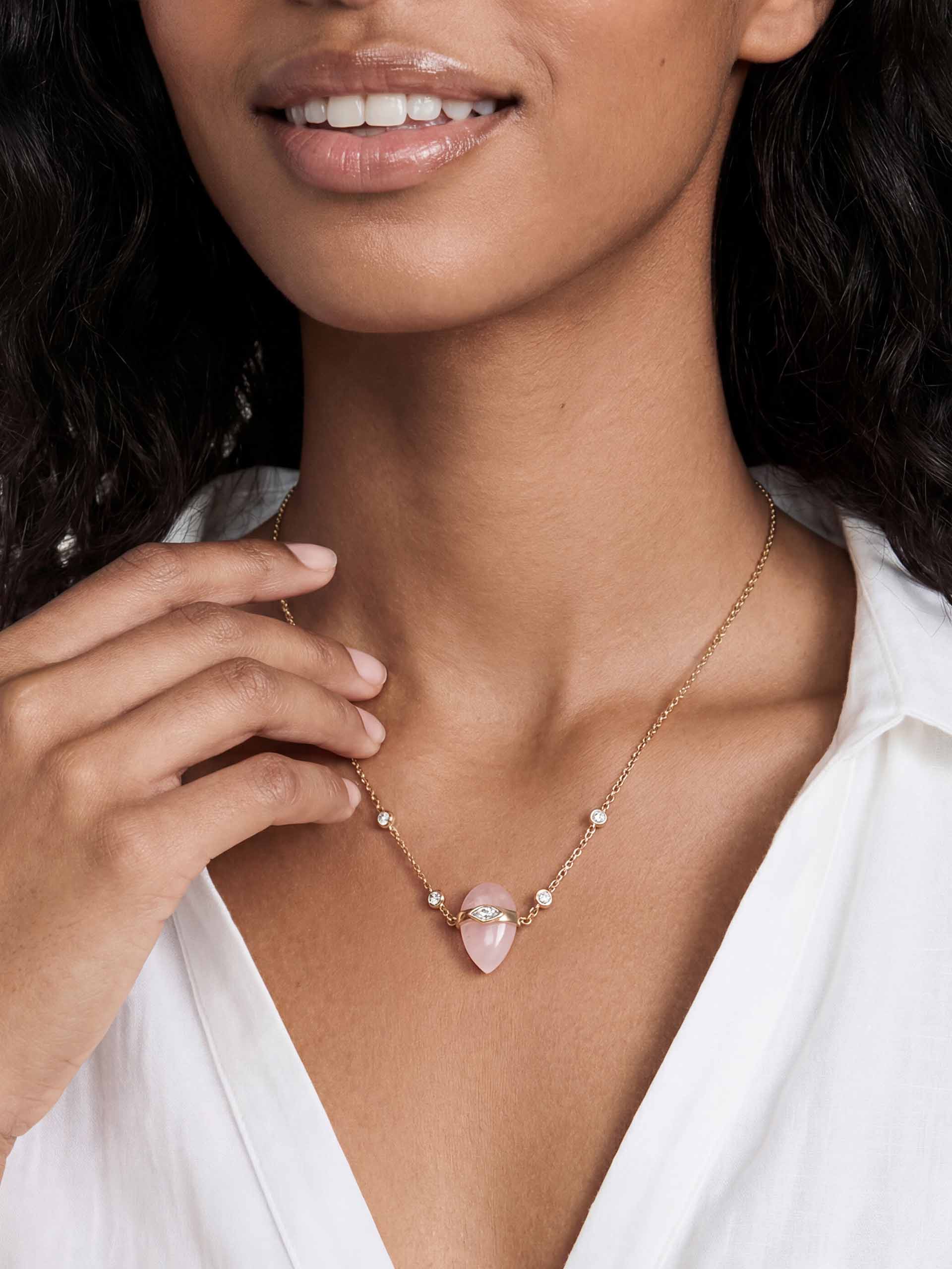 A woman wears a delicate gold necklace with small diamonds and a pink gemstone pendant, set against her bare neck. She wears a white V-neck top.
