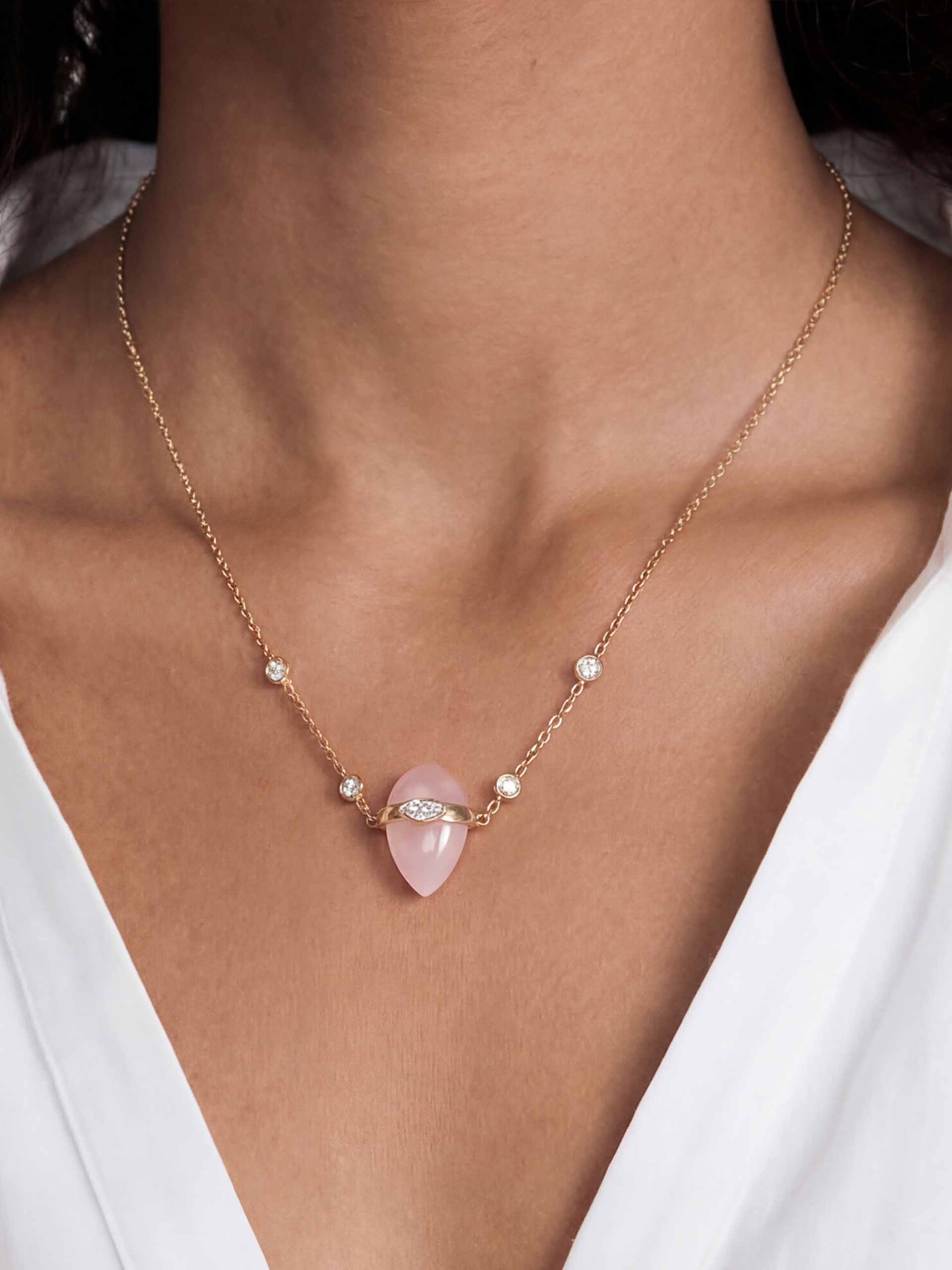 A woman wears a delicate gold necklace with small diamonds and a pink gemstone pendant, set against her bare neck. She wears a white V-neck top.