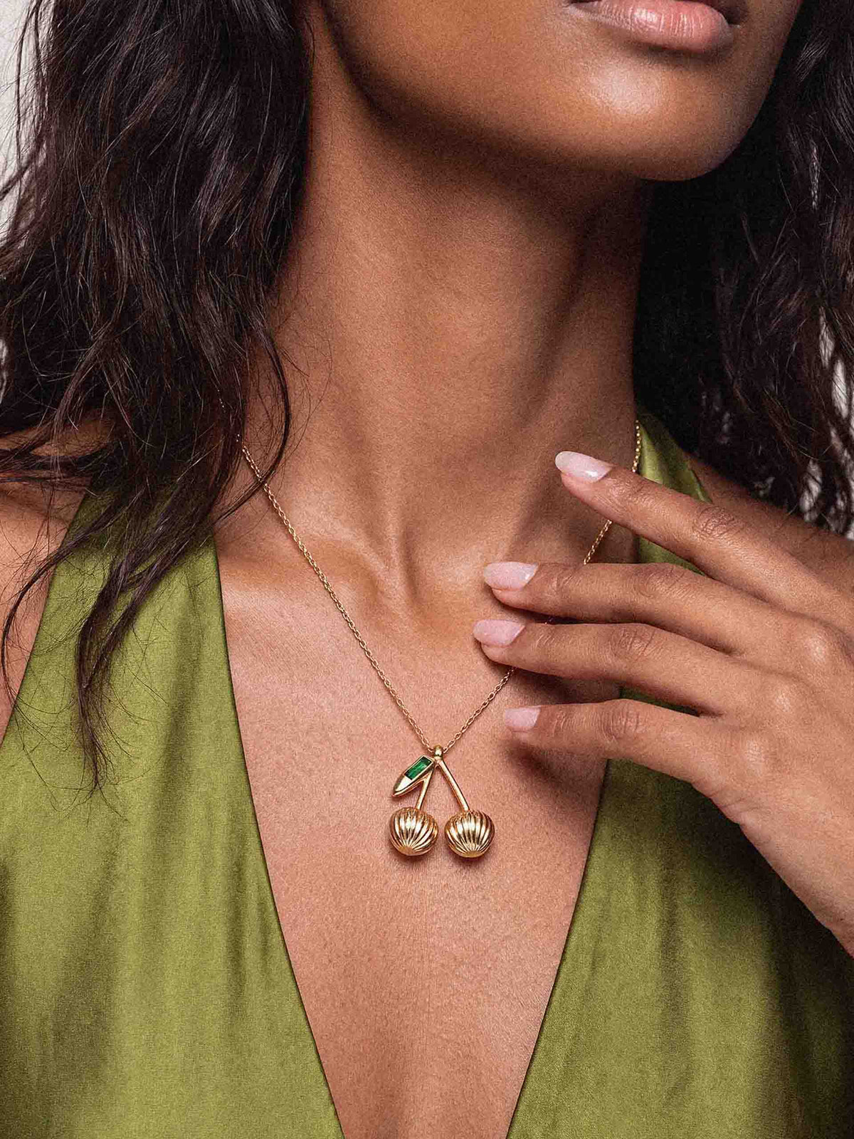 Close-up of a woman in an olive-green V-neck top, fingers near a gold necklace with twin textured charms and green stones; elegant, warm styling.