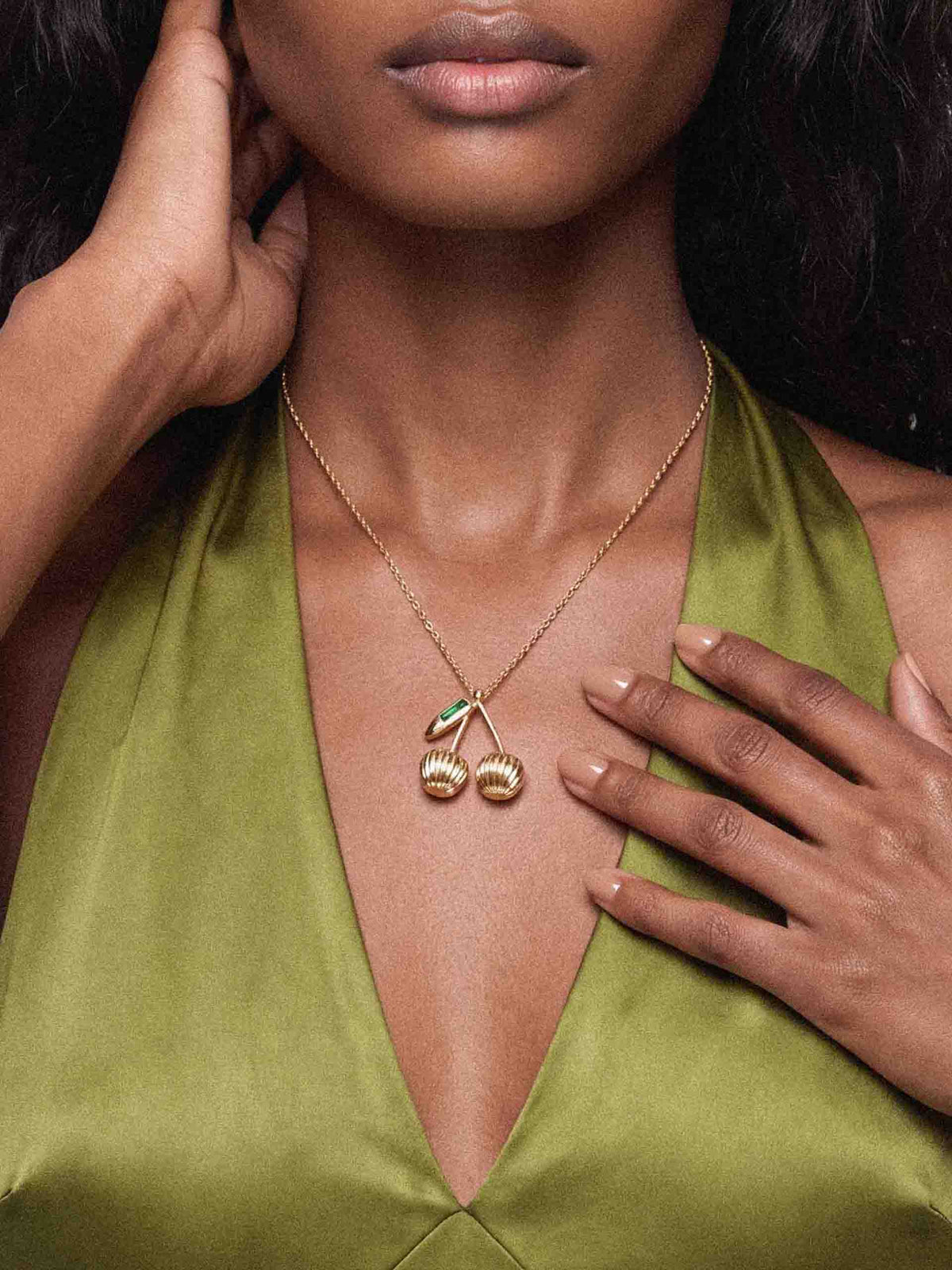 Close-up of a woman in an olive satin V-neck top, wearing a fine gold necklace with cherry charms as her hands frame her neck; elegant, poised mood.