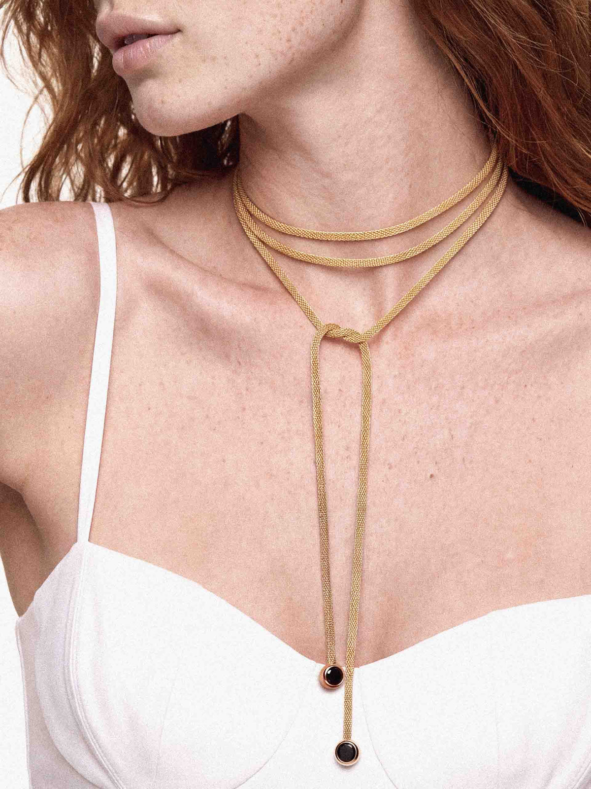 A close-up of a model wearing a multi-layered tan cord choker necklace with two small dark round beads at the end, paired with a white camisole top.
